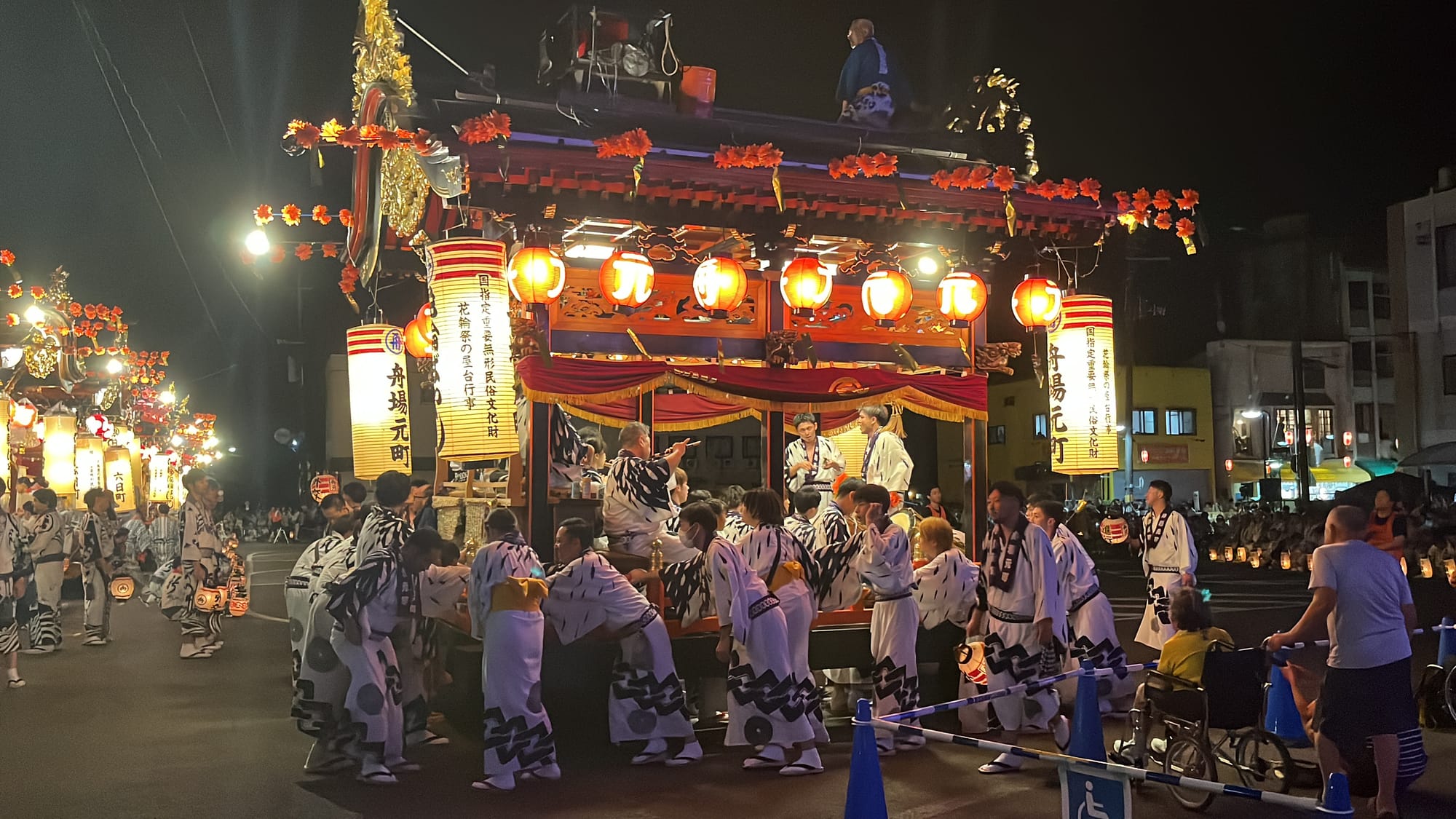 Lantern-lit festival float in Kazuno surrounded by participants in traditional dress, with spectators watching as the procession moves through the streets at night.