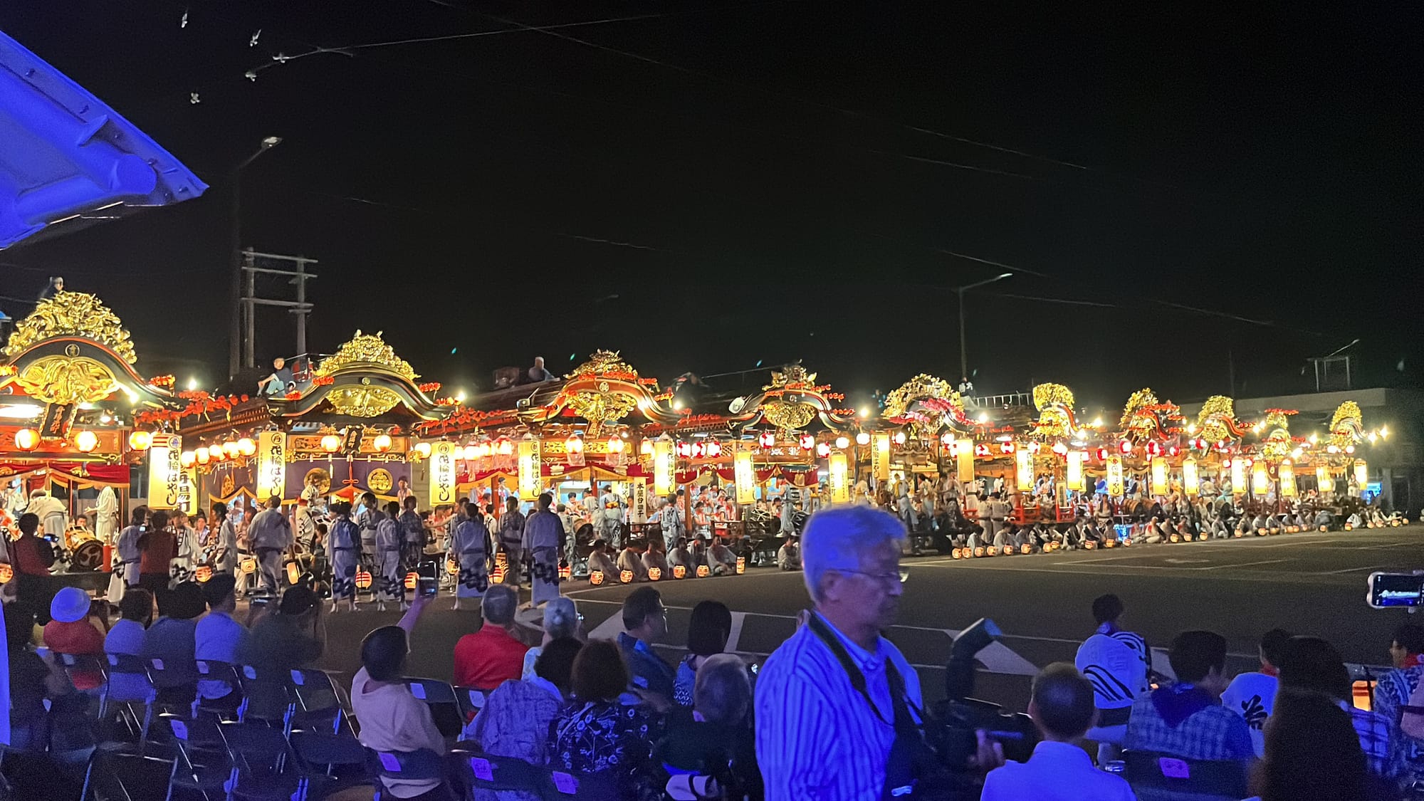 Rows of illuminated festival floats line the streets of Kazuno at night, with lanterns, musicians and spectators gathered for a traditional summer celebration.
