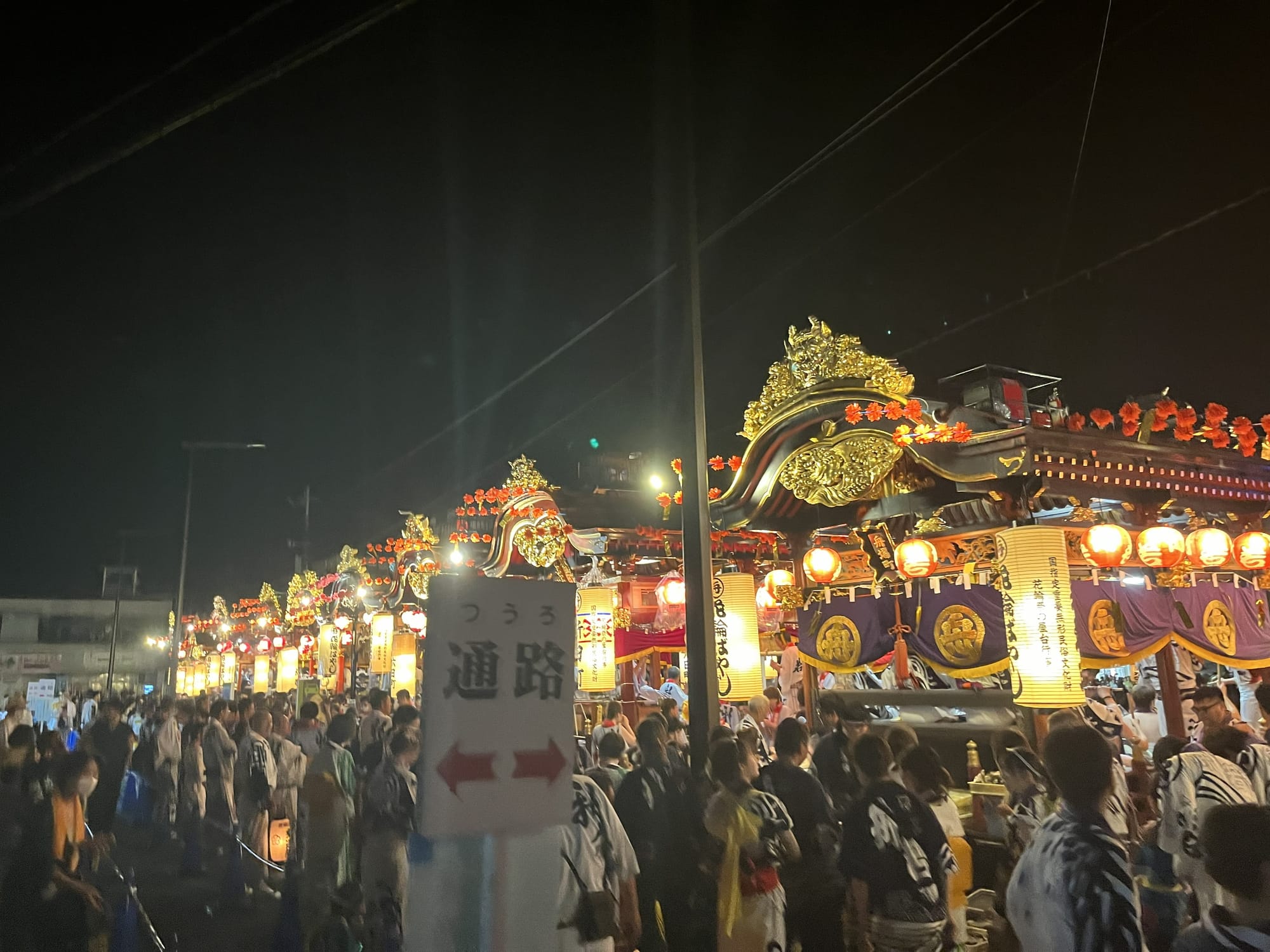 Rows of illuminated festival floats line the streets of Kazuno at night, surrounded by crowds of spectators and participants in traditional dress.