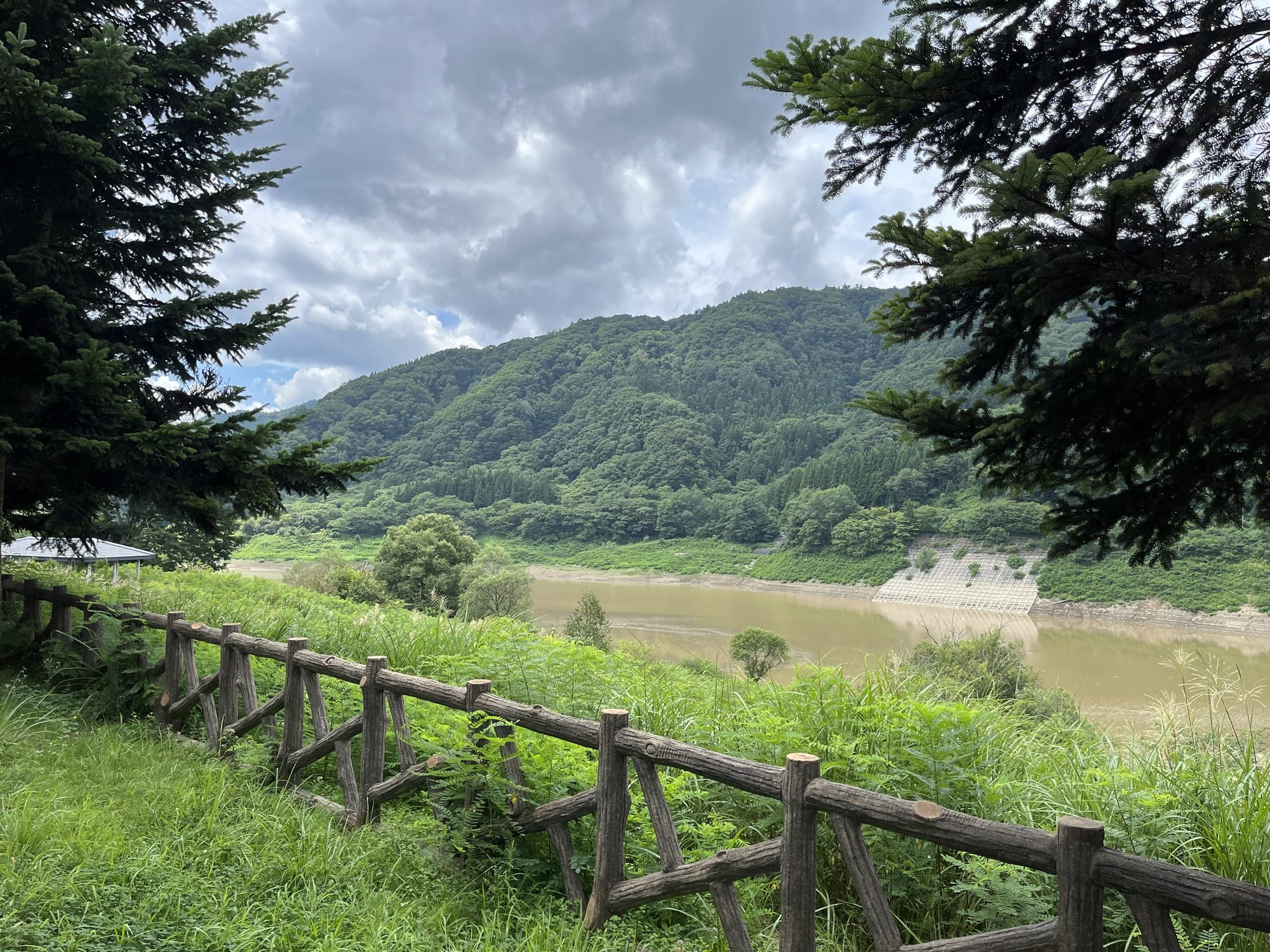View across Lake Oirase, with forested hills reflected in calm water, framed by trees and a wooden fence beneath dramatic summer clouds.