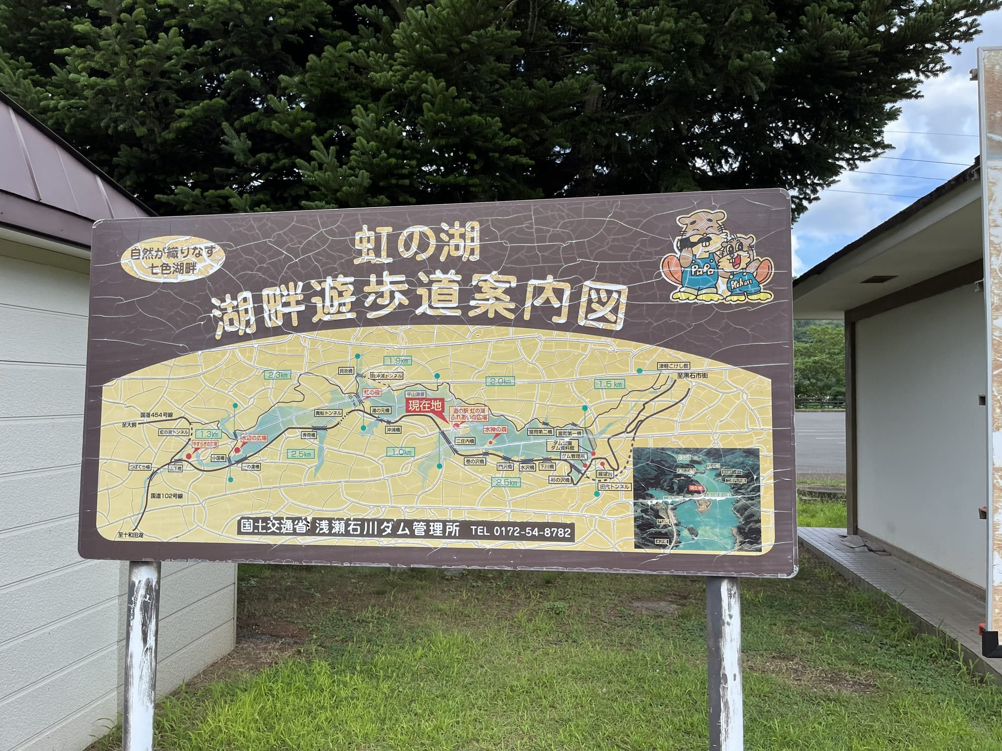 Information board showing a walking trail map around Lake Oirase, with route distances, viewpoints and local attractions marked, set against trees and nearby buildings.