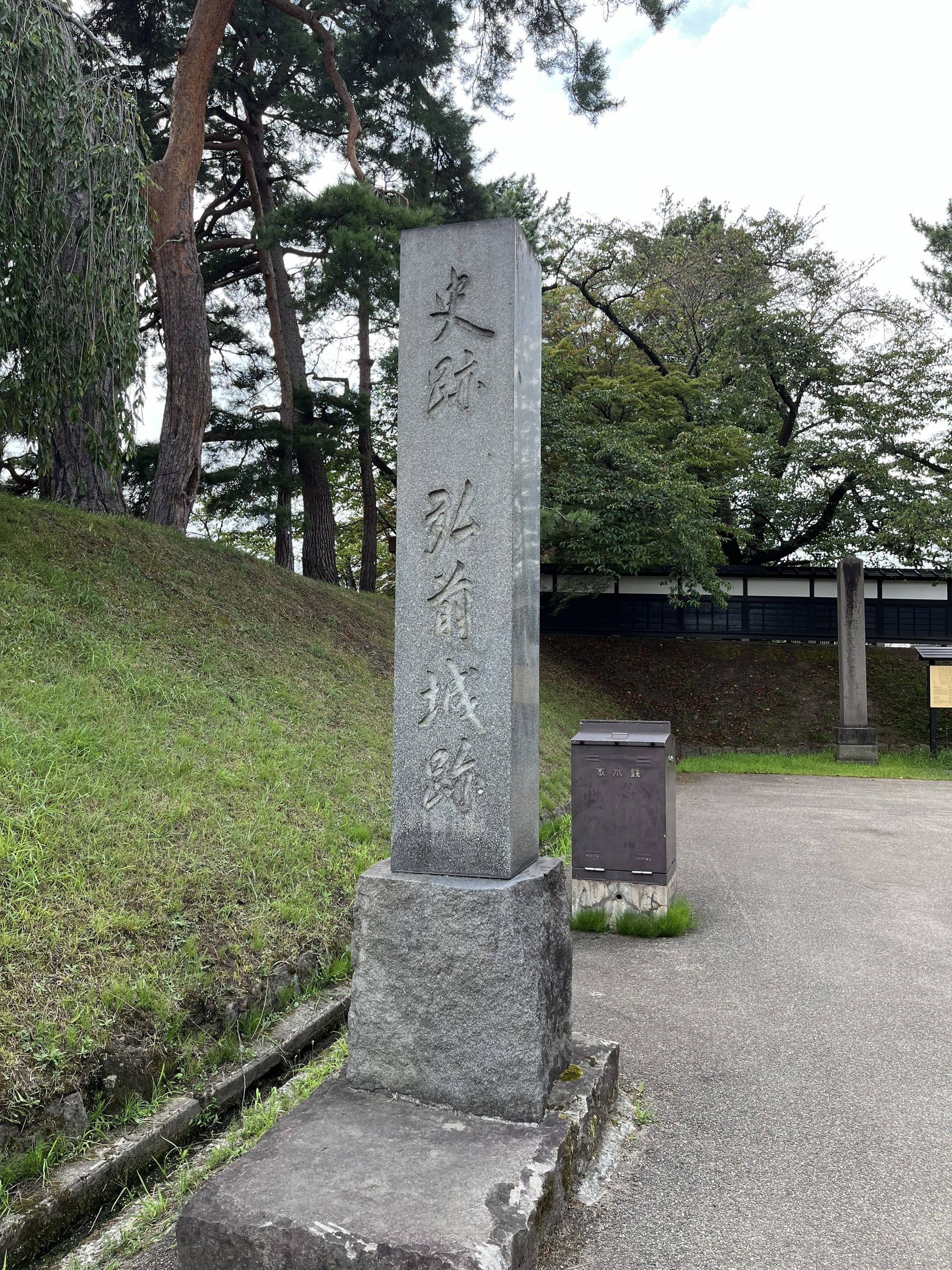 Stone monument at Hirosaki Castle bearing Japanese inscriptions, standing beside an earthen embankment, pine trees and historic castle grounds.