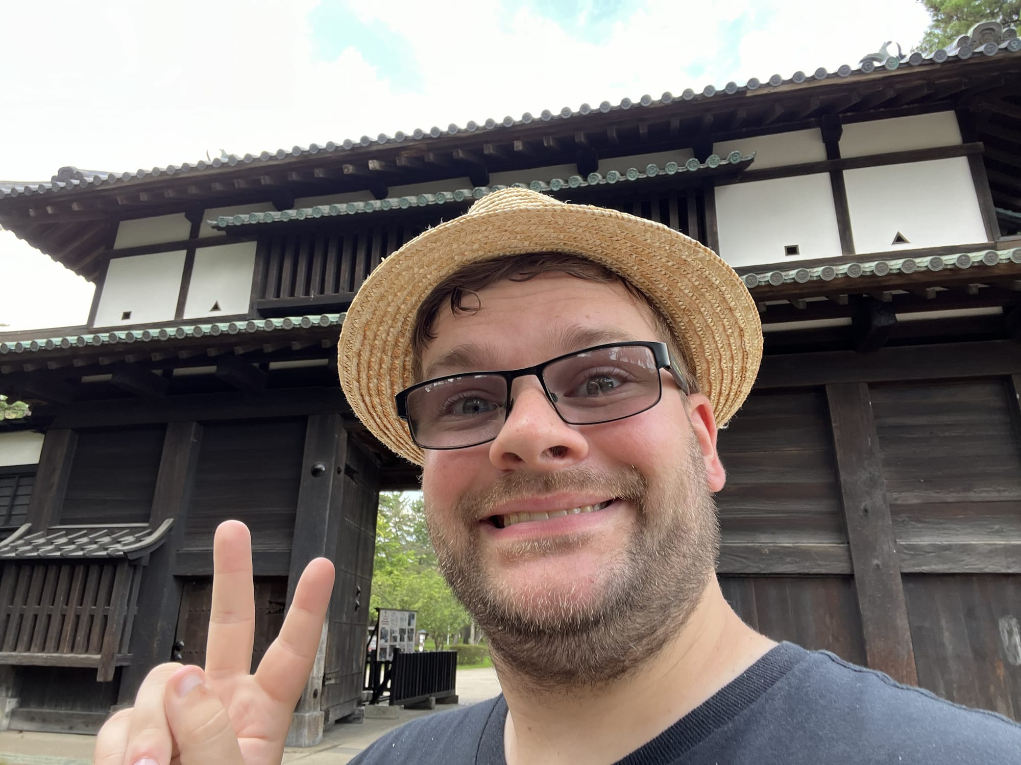 Visitor making a peace sign in front of a traditional gate at Hirosaki Castle, with dark wooden architecture and tiled roofs visible in the background.