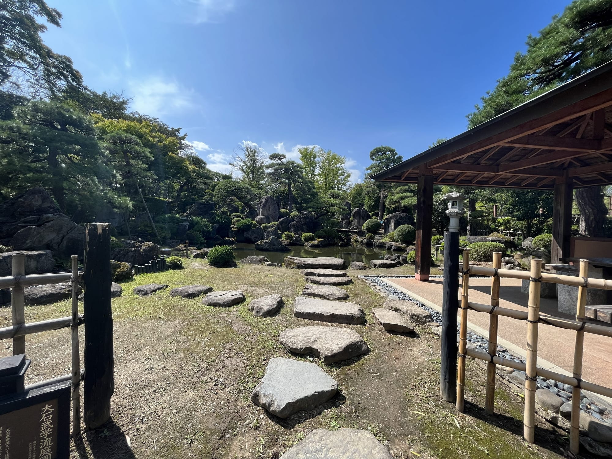 Traditional Japanese garden at Hirosaki Castle, featuring stepping stones, a pond, carefully pruned trees, rock formations and a wooden pavilion beneath a clear blue sky.
