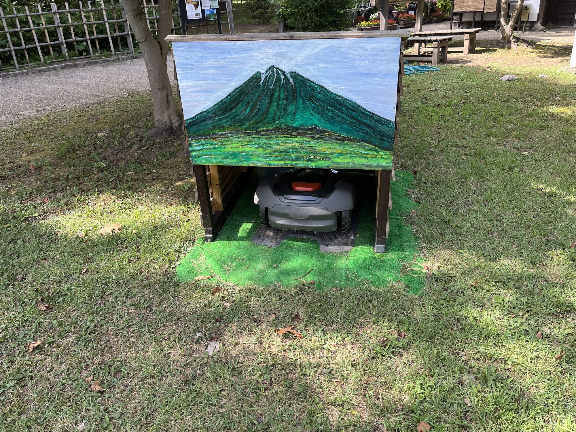 Robotic lawn mower parked beneath a painted wooden shelter in the Hirosaki Castle Botanical Gardens, with artwork depicting Mount Iwaki on the front panel.