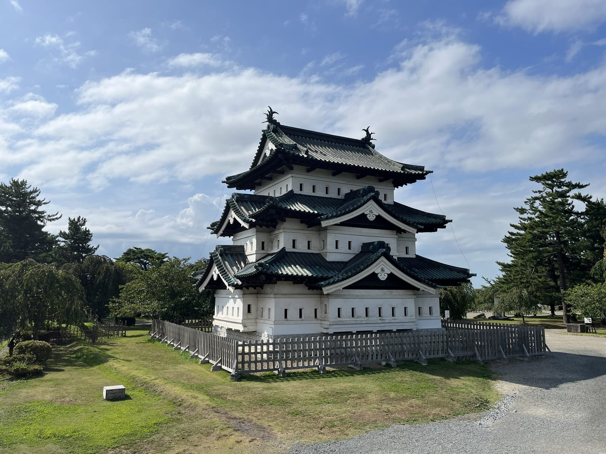 Hirosaki Castle keep in Aomori Prefecture, featuring white walls and dark tiled roofs, surrounded by lawns and trees beneath a bright blue sky with scattered clouds.