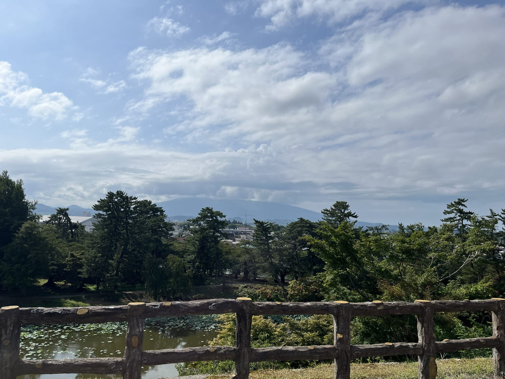 View from Hirosaki Castle across a pond and treetops towards Mount Iwaki, with dramatic clouds gathering above the mountain beneath a bright summer sky.