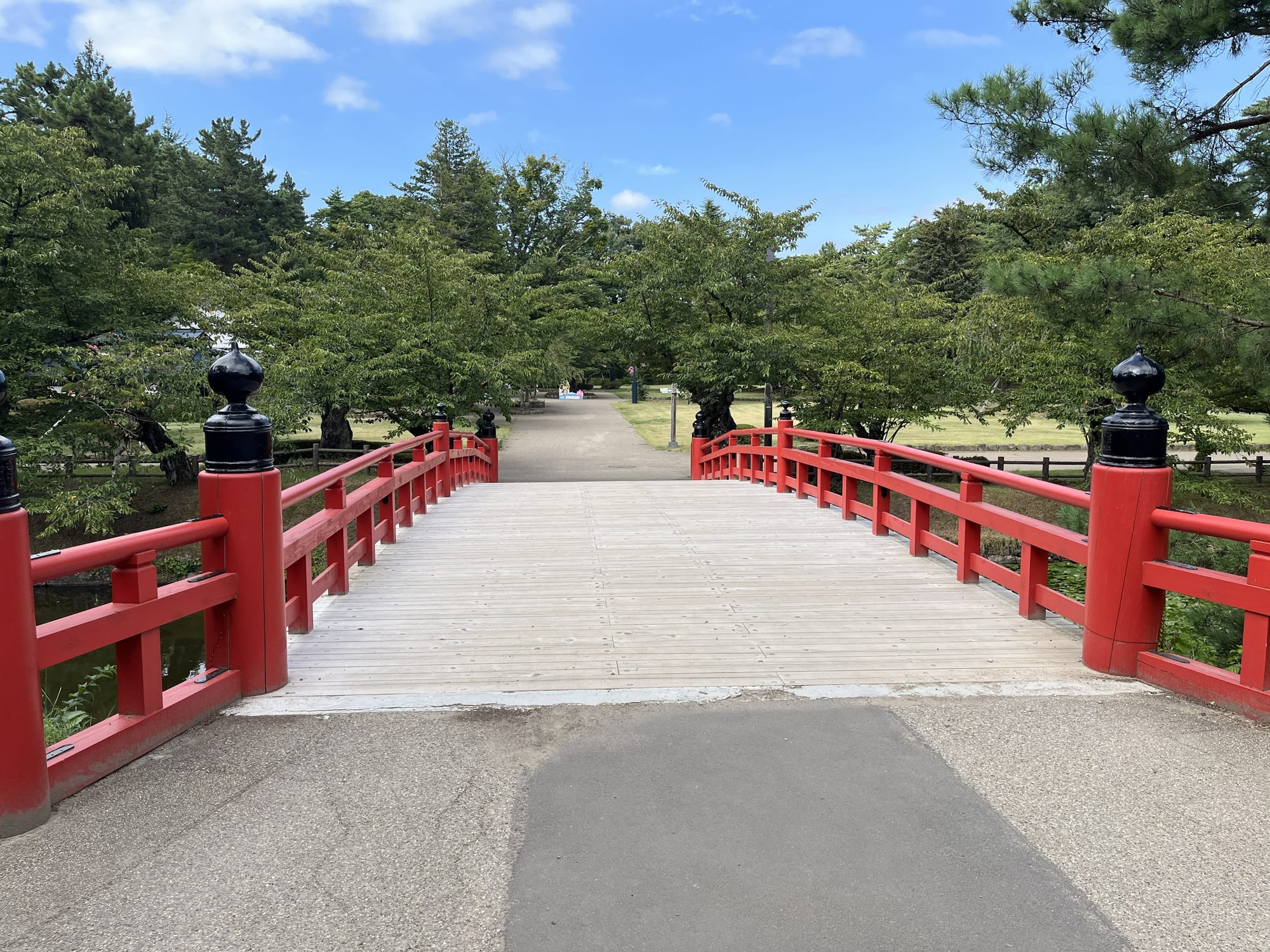 Red wooden bridge at Hirosaki Castle crossing a moat, with trees and landscaped grounds beyond, beneath a bright blue sky with scattered clouds.