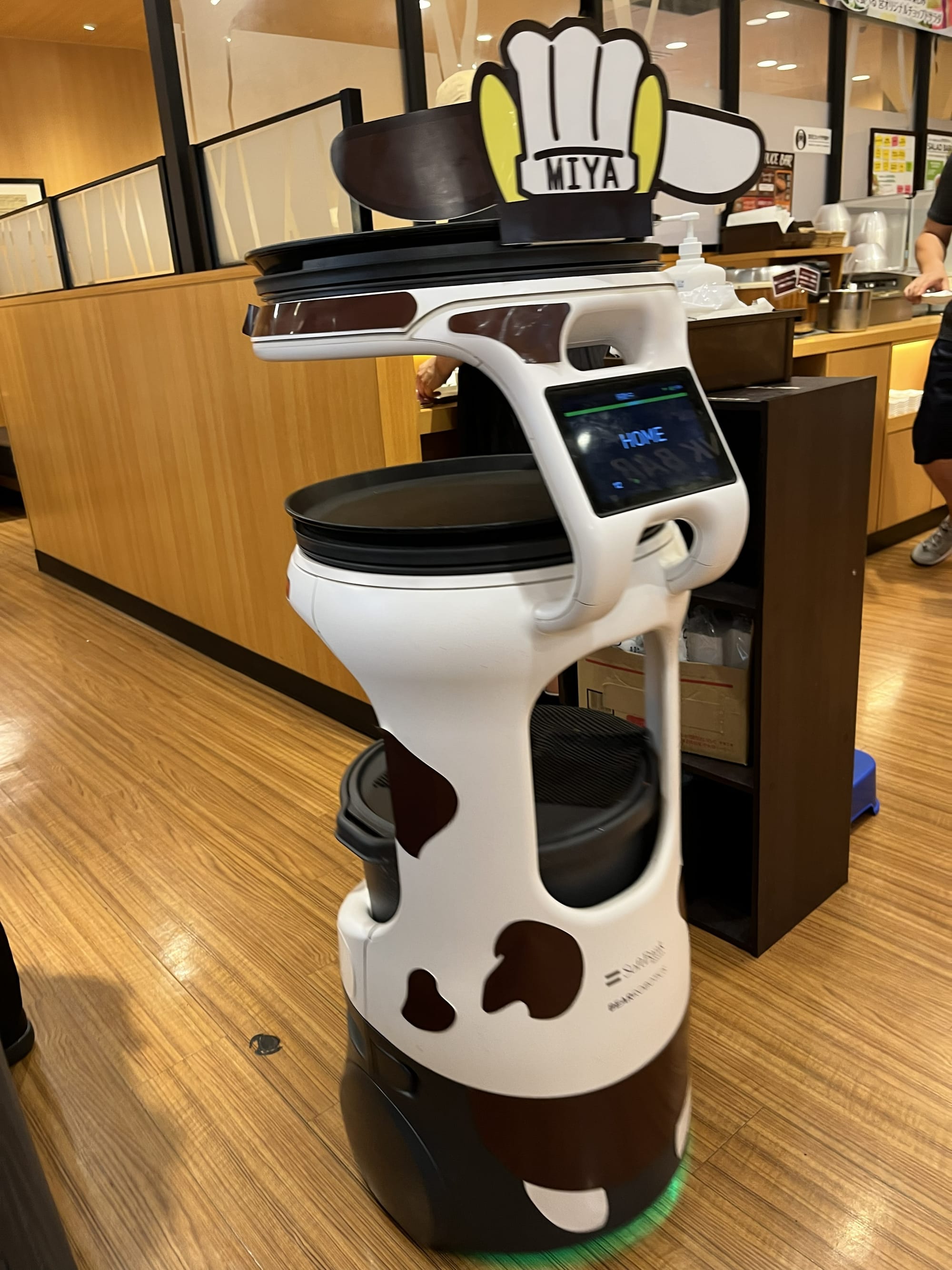 Cow-themed serving robot in a Japanese family restaurant, with trays for carrying food and a digital display, standing beside booths and wooden furnishings.
