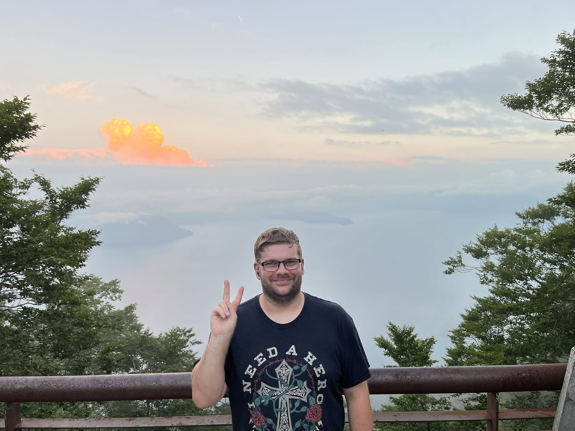 Person standing at a lakeside viewpoint above Lake Towada, with misty water, forested shores and orange sunset clouds visible beyond the railing in northern Japan.
