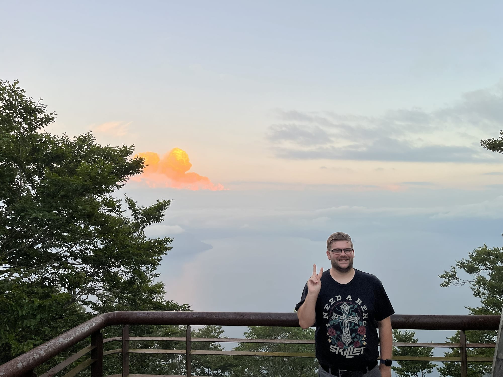 Person standing at a viewpoint above Lake Towada, making a peace sign as sunset clouds glow orange over the misty lake and forested shoreline in northern Japan.