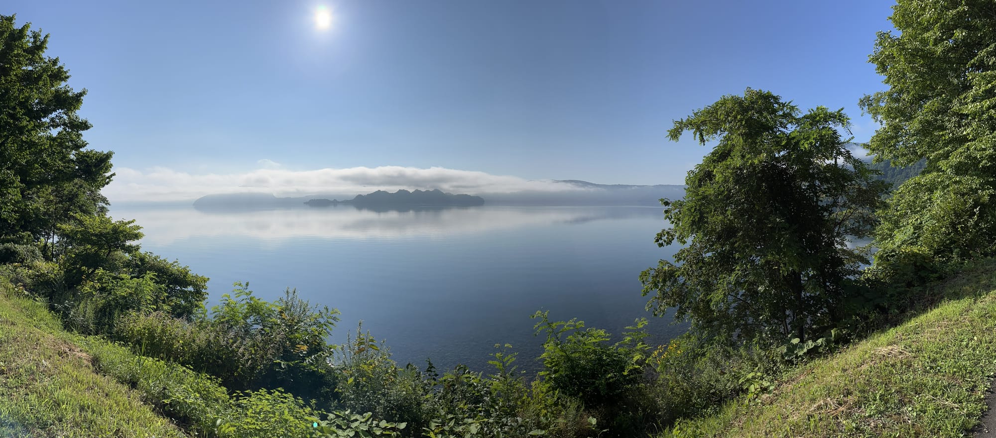 View across Lake Towada framed by lakeside trees, with calm reflective water, low cloud over the distant shoreline and bright morning sunshine above the caldera lake.