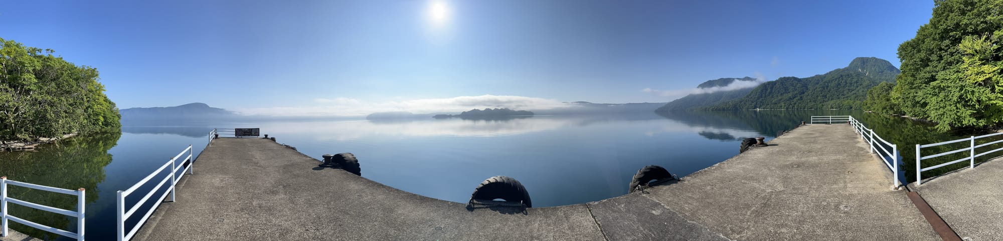 Panoramic view from a pier on Lake Towada, with calm water reflecting forested mountains, light mist above the shoreline and clear blue skies overhead.