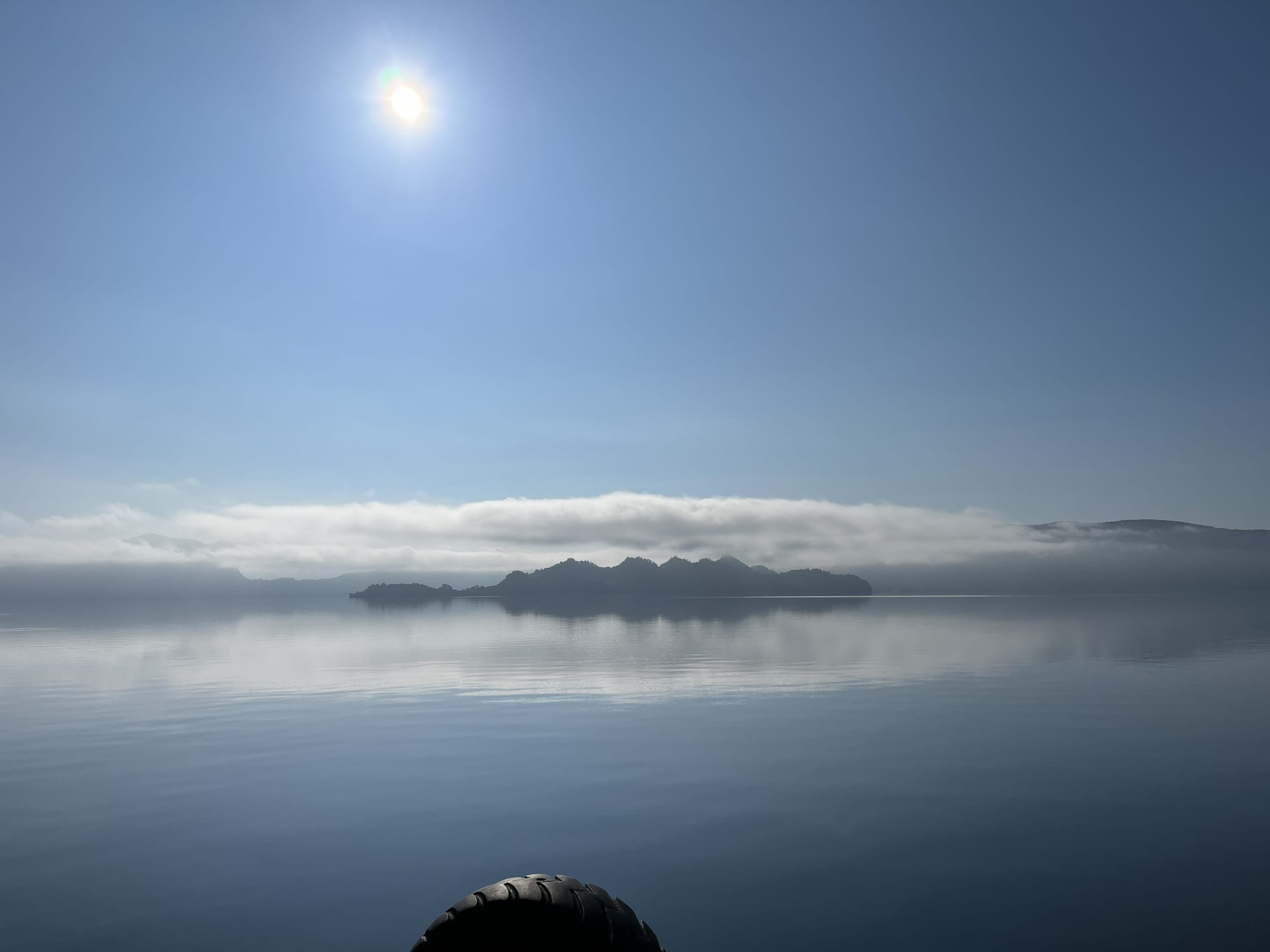 Calm waters of Lake Towada reflecting low cloud and blue sky, with the silhouette of Nakayama Peninsula visible across the lake beneath the bright morning sun.