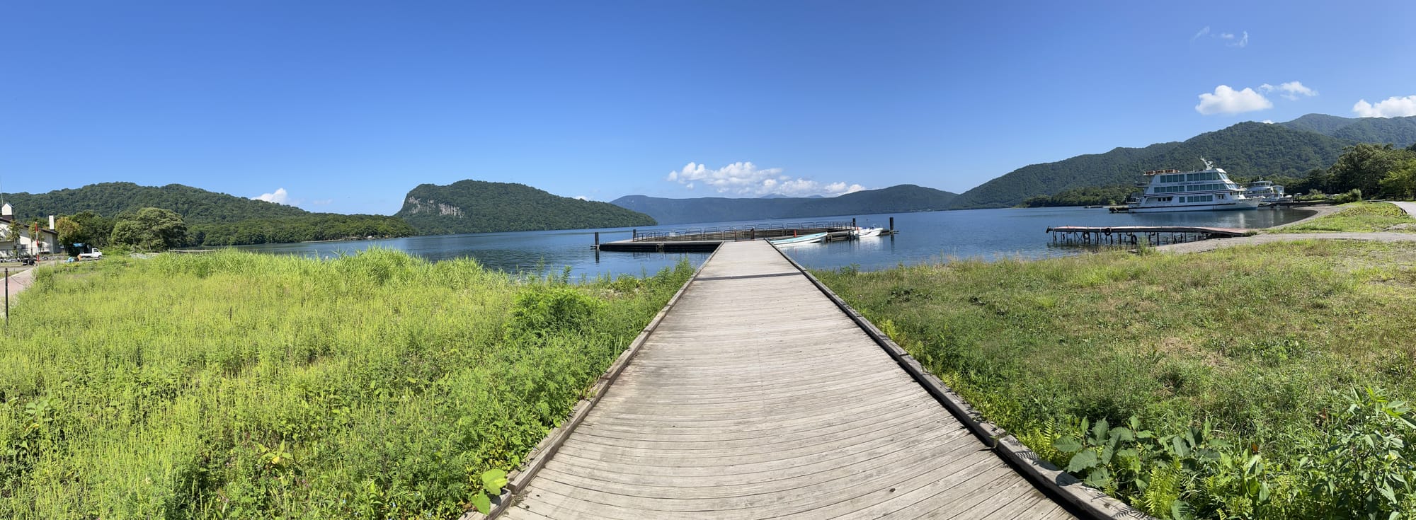 Wooden boardwalk leading to a pier on Lake Towada, with calm blue water, forested mountains, a sightseeing boat and clear skies on a bright summer day.