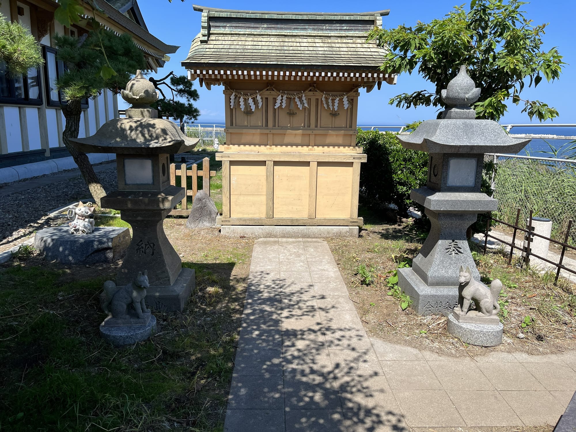 Small wooden shrine at Kabushima Shrine, flanked by stone lanterns and fox statues, with coastal views and greenery surrounding the sacred site in Hachinohe.