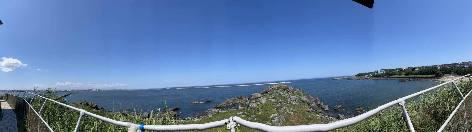 View from Kabushima overlooking rocky coastline and the Pacific Ocean, with grassy cliffs, breakwaters, coastal buildings and clear blue skies stretching across Hachinohe Bay.