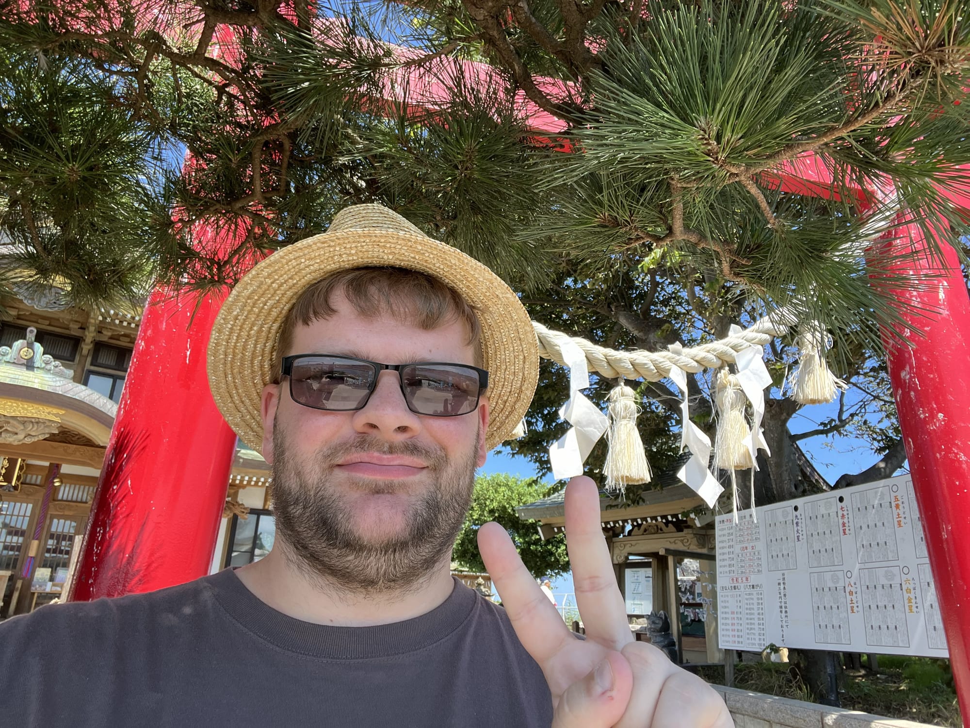 Visitor wearing a straw hat and sunglasses stands beneath a red torii gate at Kabushima Shrine, with sacred shimenawa rope, pine branches and shrine buildings behind.