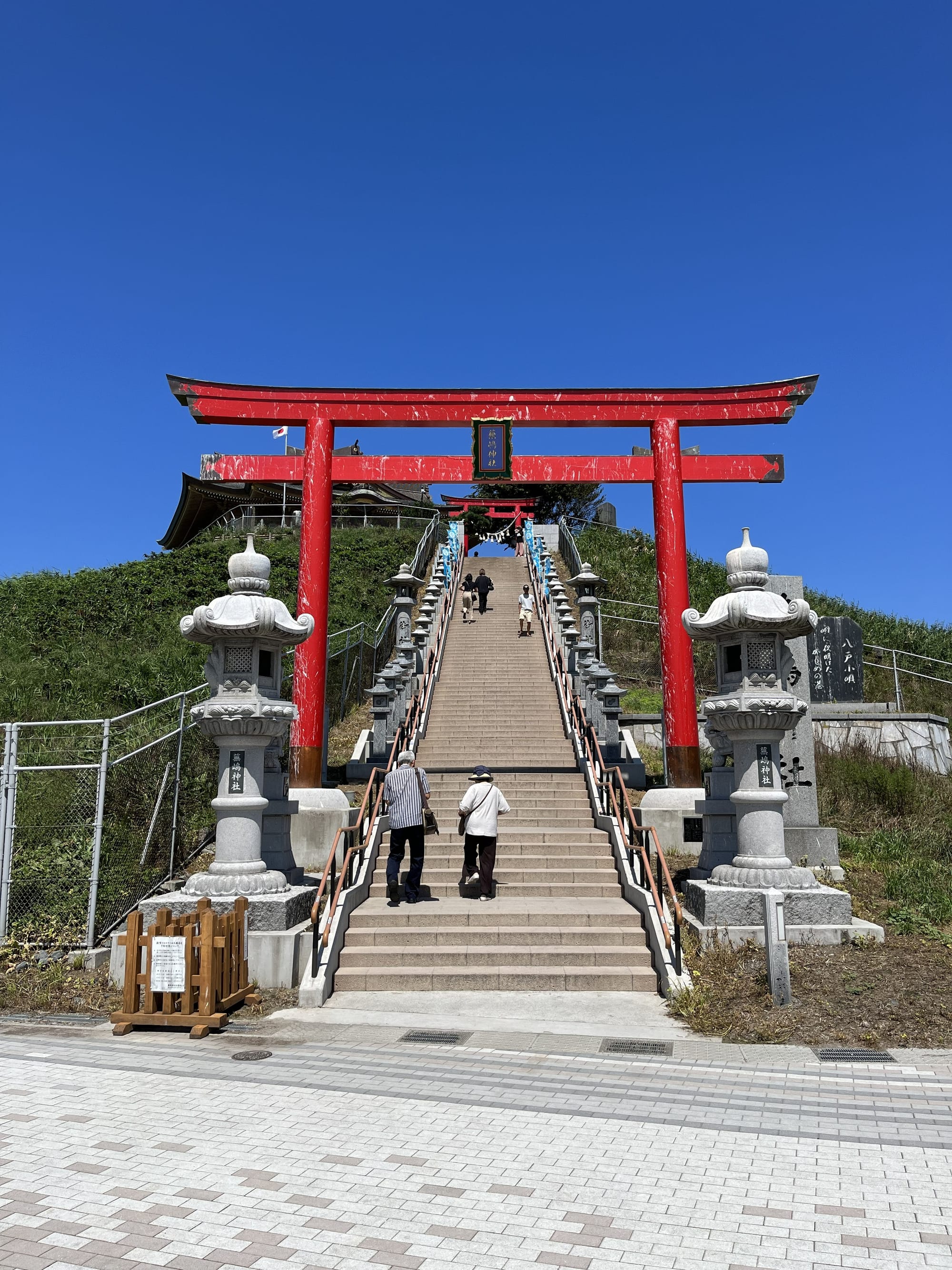 Large red torii gate and stone lanterns at the entrance to Kabushima Shrine, with visitors climbing a long staircase towards the hilltop shrine under clear blue skies.