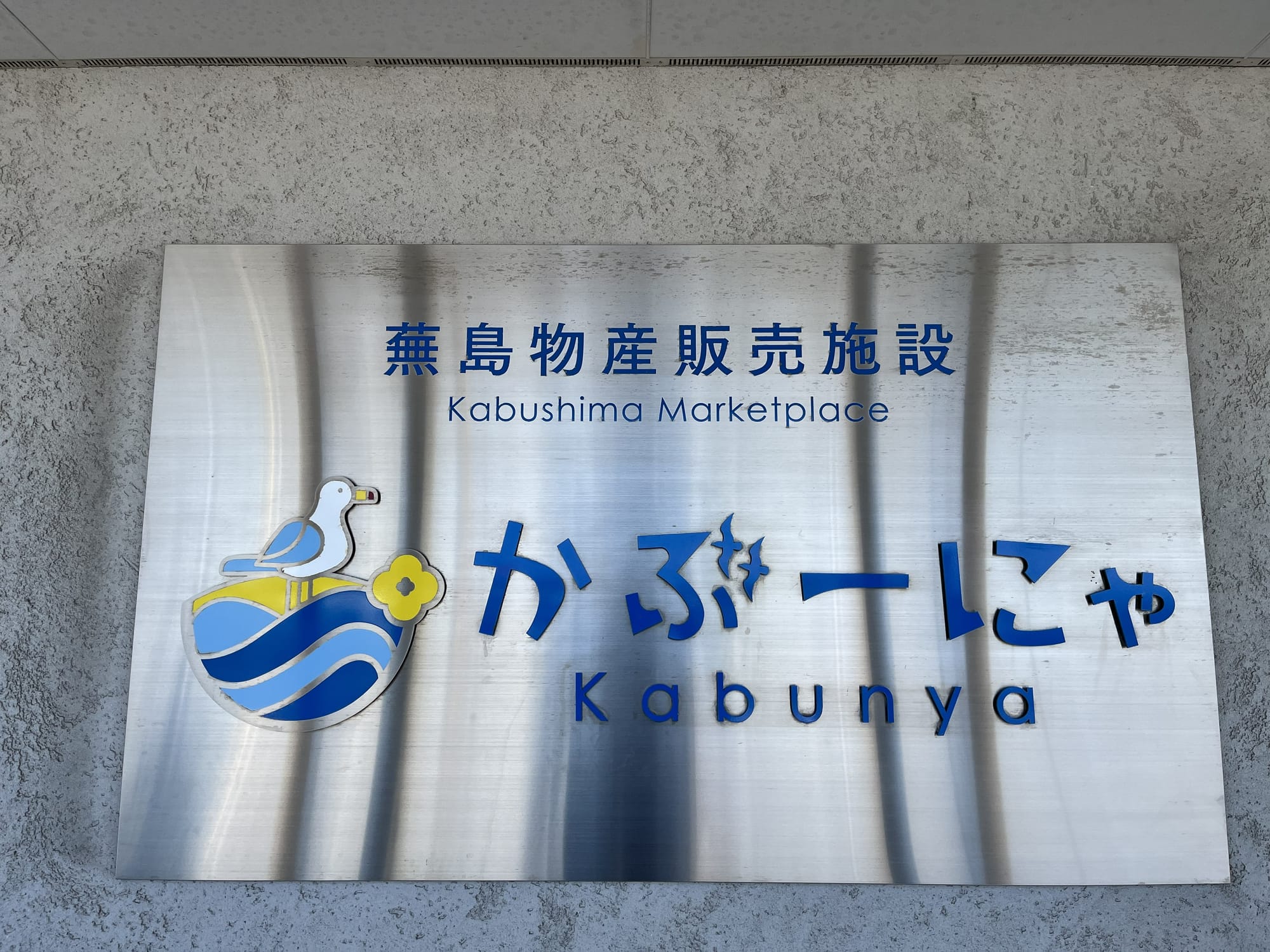 Metal sign for Kabunya at Kabushima Marketplace, featuring Japanese and English text alongside a seagull logo in blue, white and yellow.