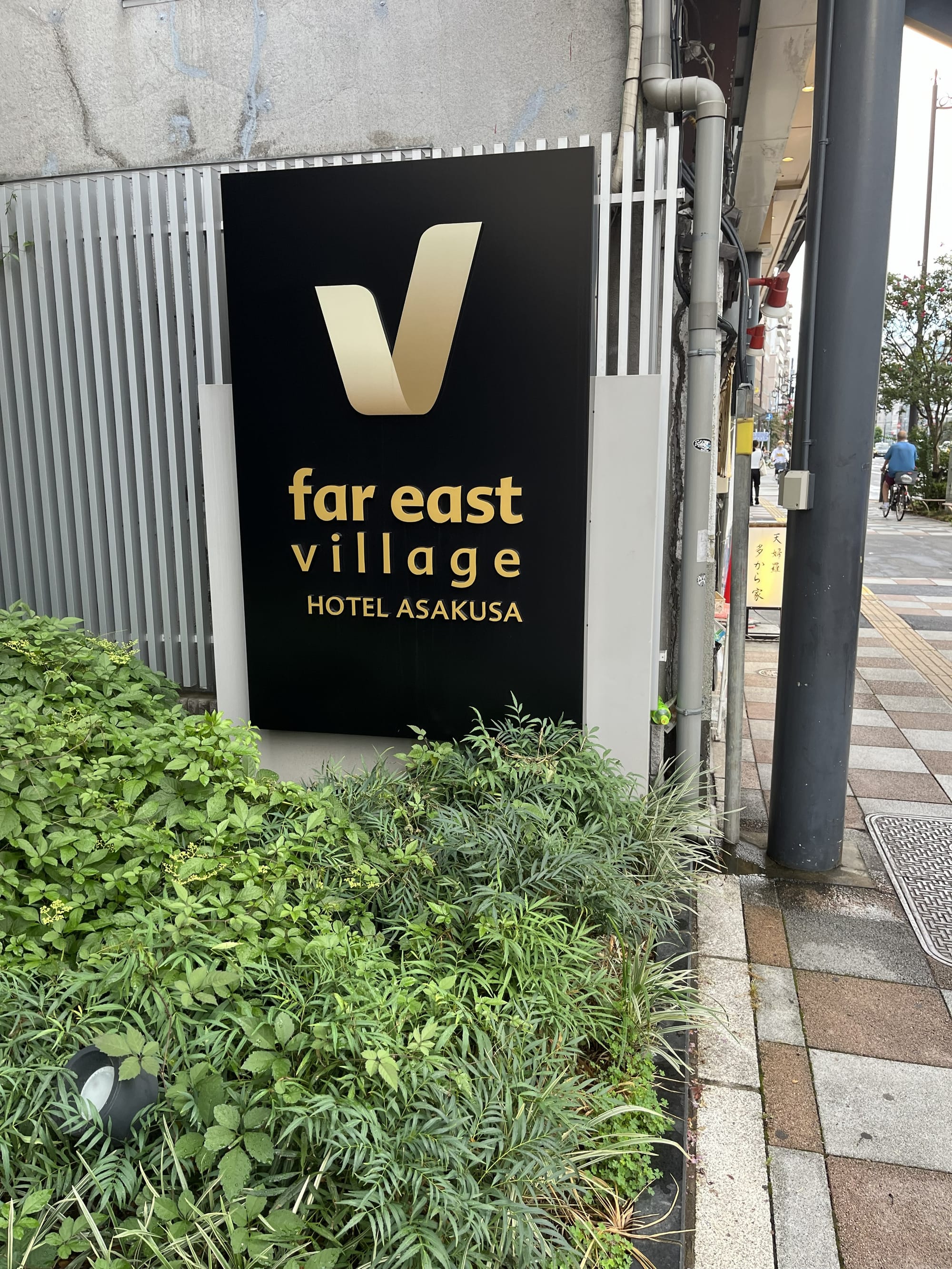 Sign for Far East Village Hotel Asakusa beside a Tokyo street, featuring gold lettering on a black background with landscaped greenery in front.