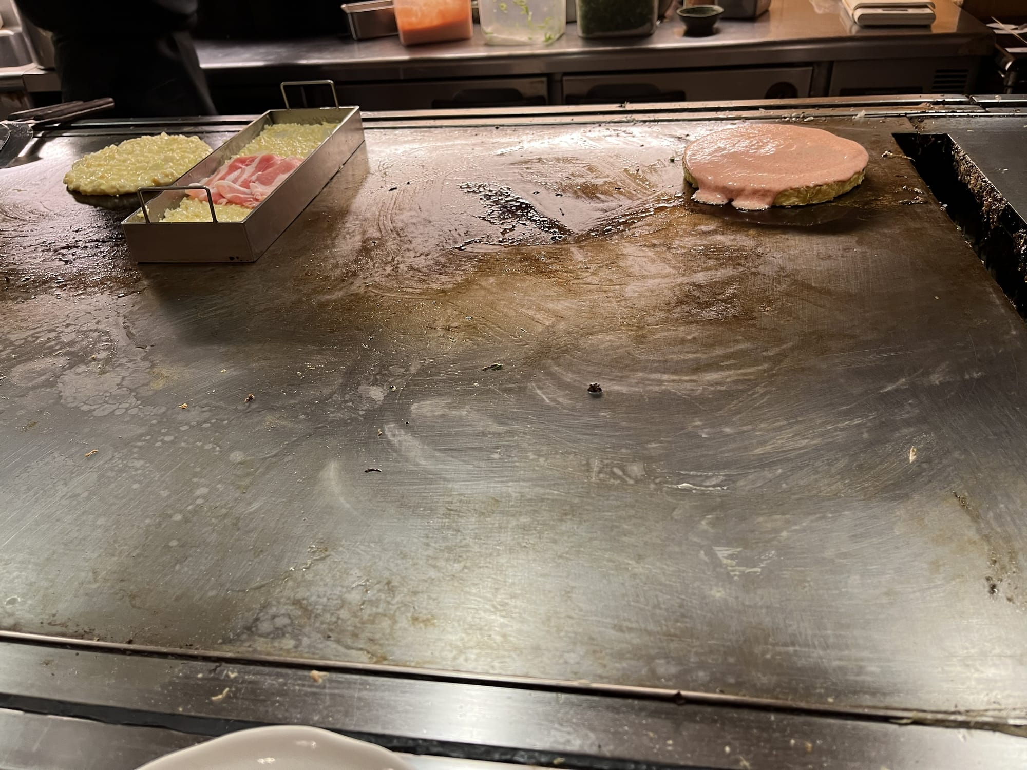 Okonomiyaki cooking on a hot teppan grill, with batter, cabbage and fillings being prepared by a chef in an open kitchen at a Japanese restaurant.