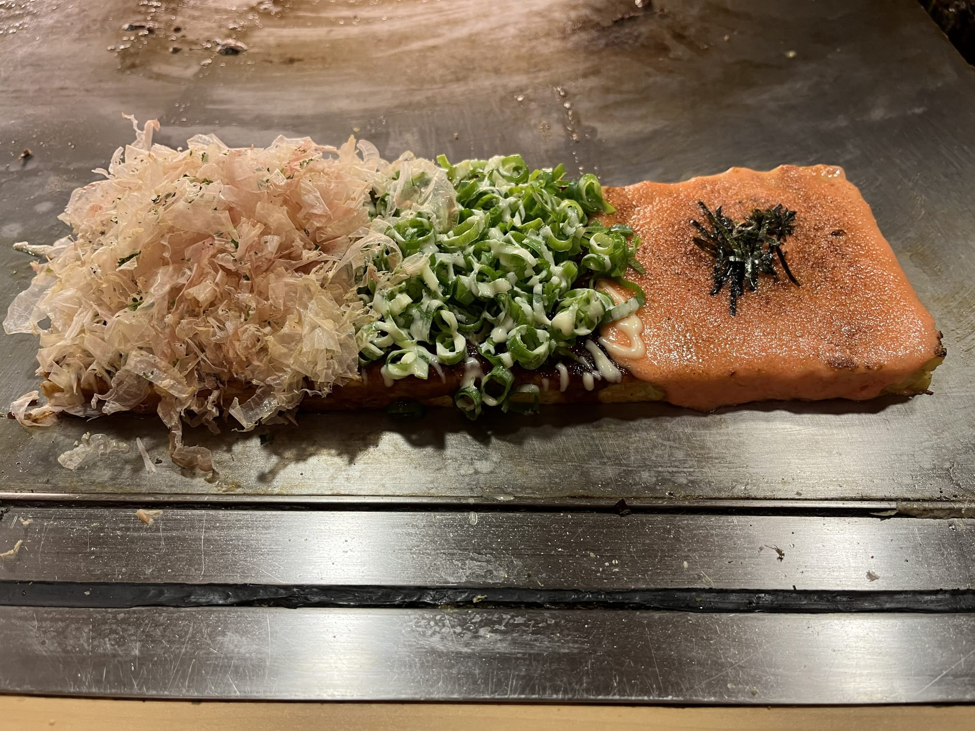 Okonomiyaki on a teppan grill topped with bonito flakes, sliced spring onions, mayonnaise and cod roe sauce, freshly cooked at a Japanese restaurant.