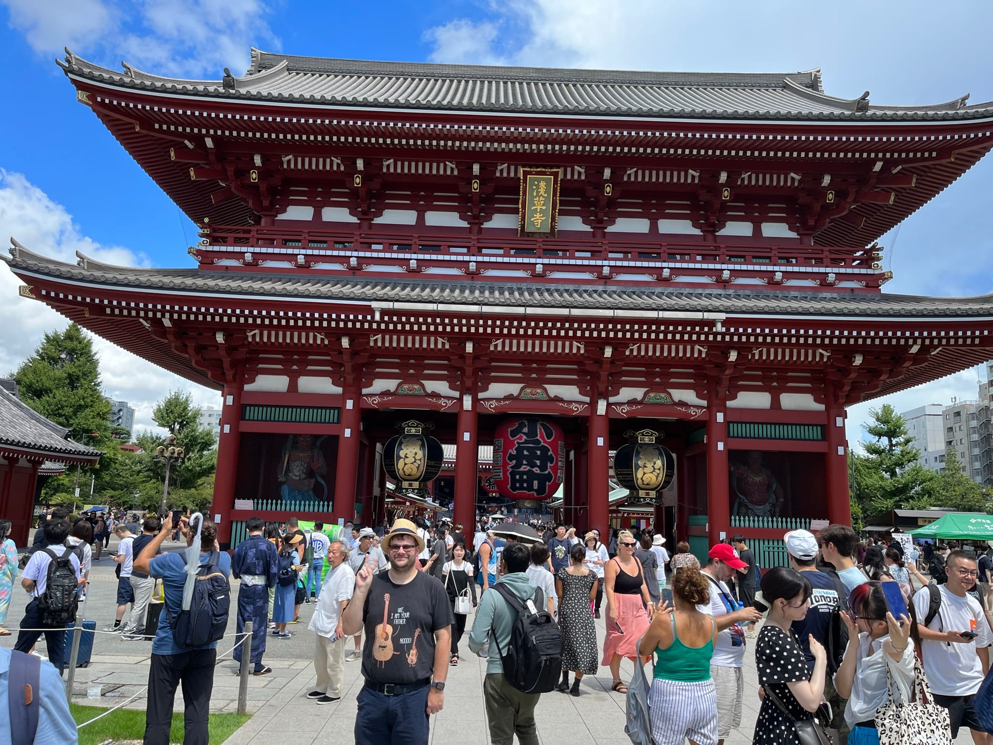 Crowds gather in front of Hōzōmon Gate at Sensō-ji Temple in Asakusa, with its large red lantern, ornate architecture and busy temple precinct on a sunny day.