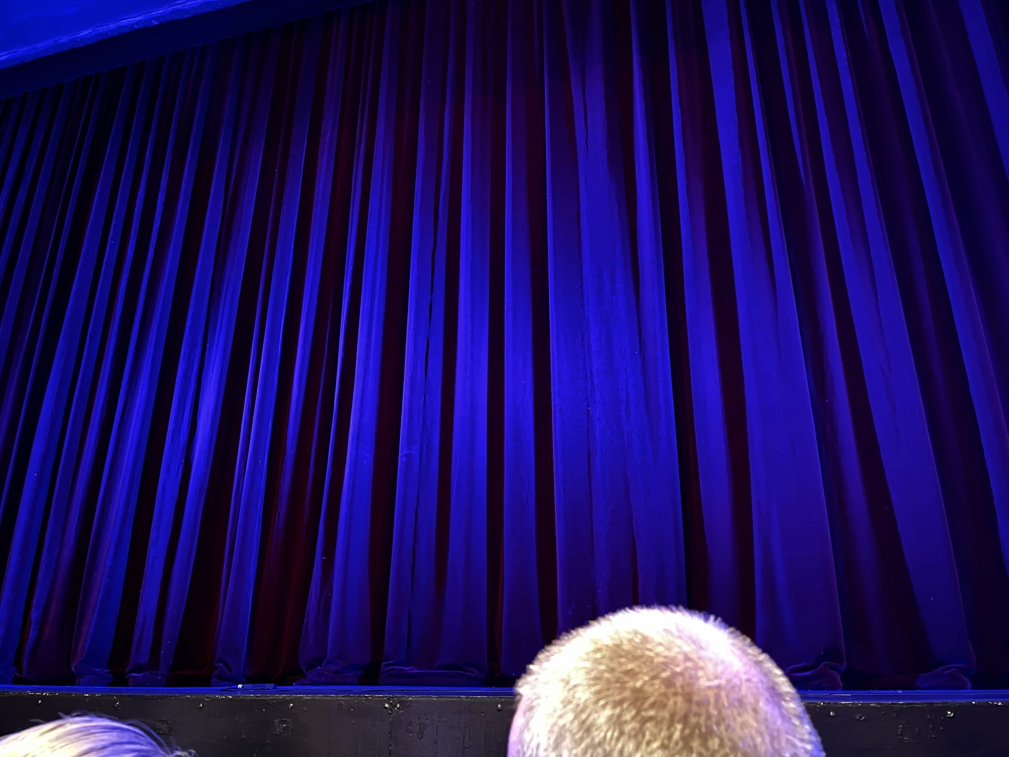 Blue stage curtain at the Peacock Theatre in London moments before a performance, illuminated by blue and purple theatrical lighting, with the heads of audience members visible in the foreground.