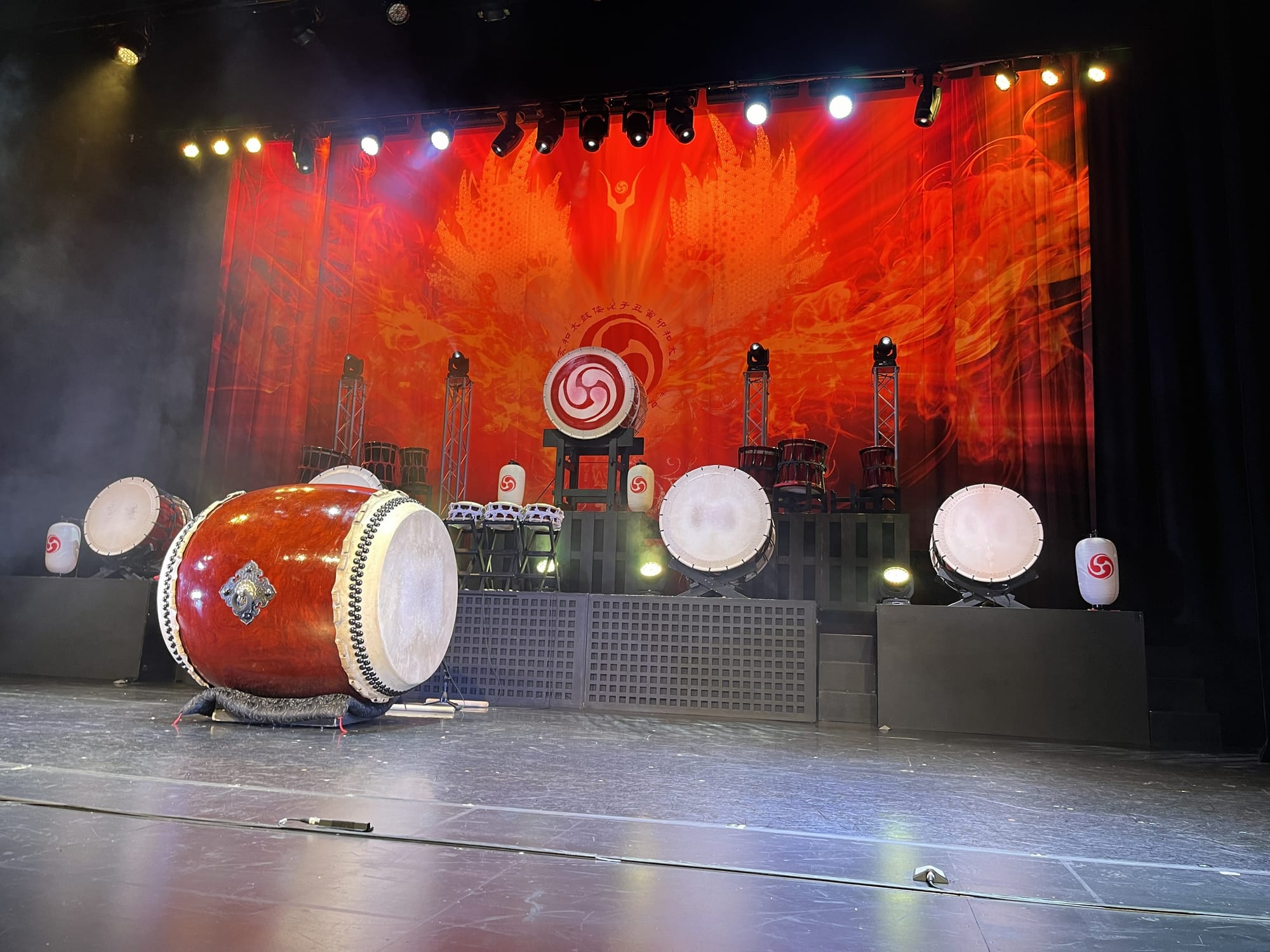 Stage set for Yamato: The Drummers of Japan at the Peacock Theatre in London, featuring a large taiko drum in the foreground, multiple drums arranged across raised platforms, dramatic red-and-orange backdrop artwork and theatrical lighting.