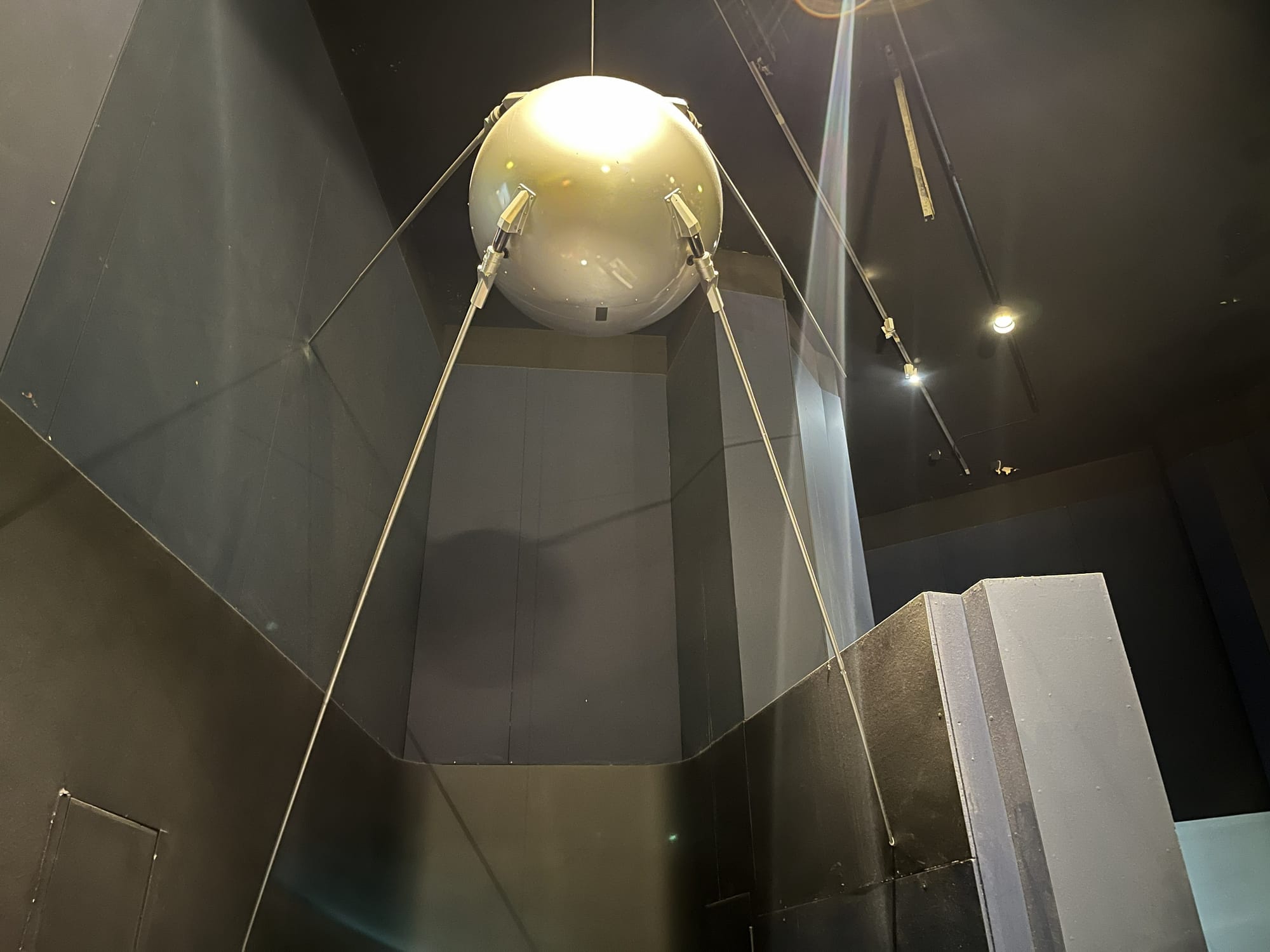 Replica of the Sputnik 1 satellite suspended in a museum gallery, featuring a polished spherical body with four long antennae extending from its surface, displayed against dark exhibition walls and illuminated by overhead lighting.