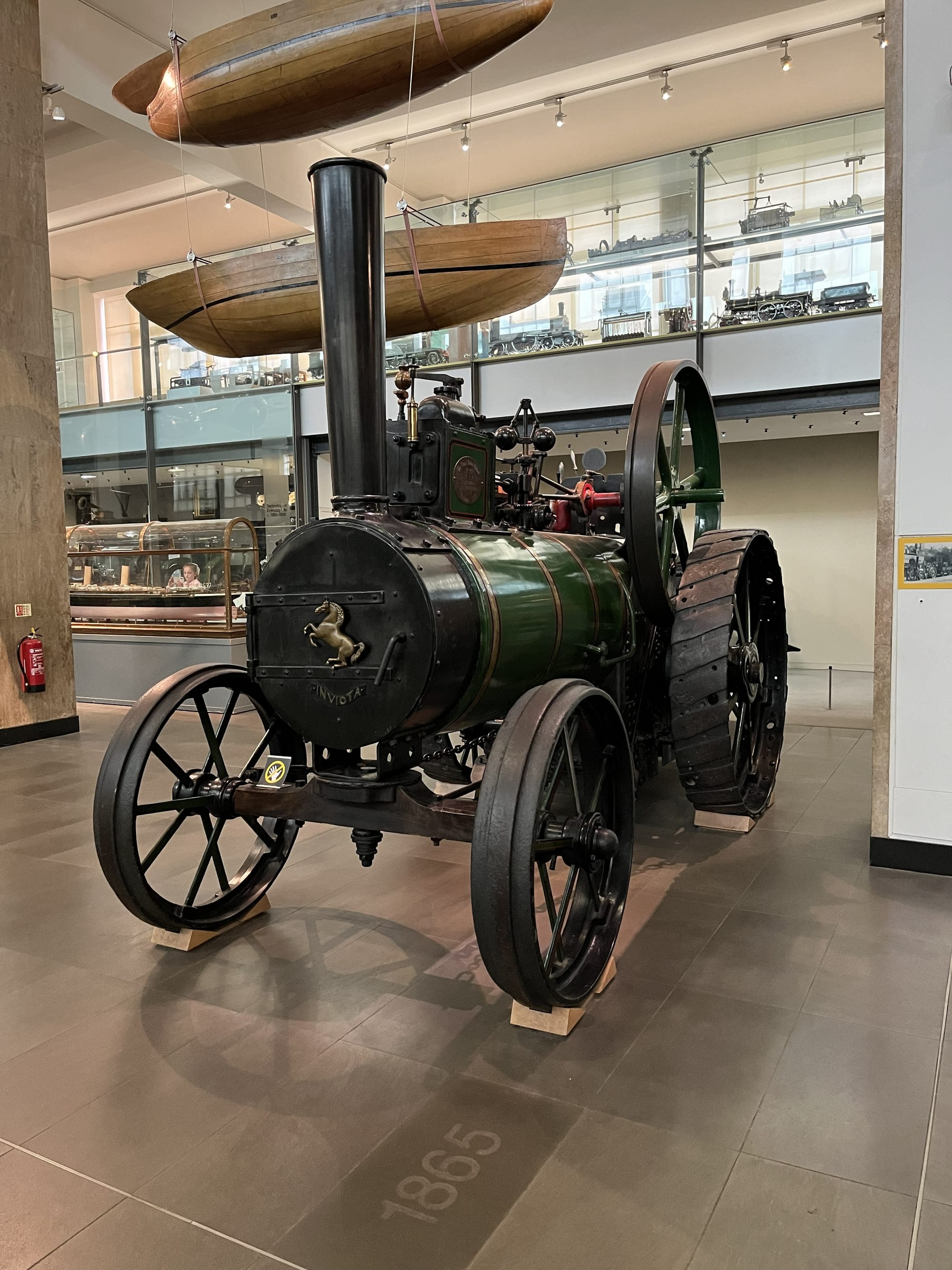 Historic steam traction engine on display at the Science Museum in London, featuring large iron wheels, a tall chimney and a green-and-black body, with suspended boat hulls and engineering exhibits visible in the background.