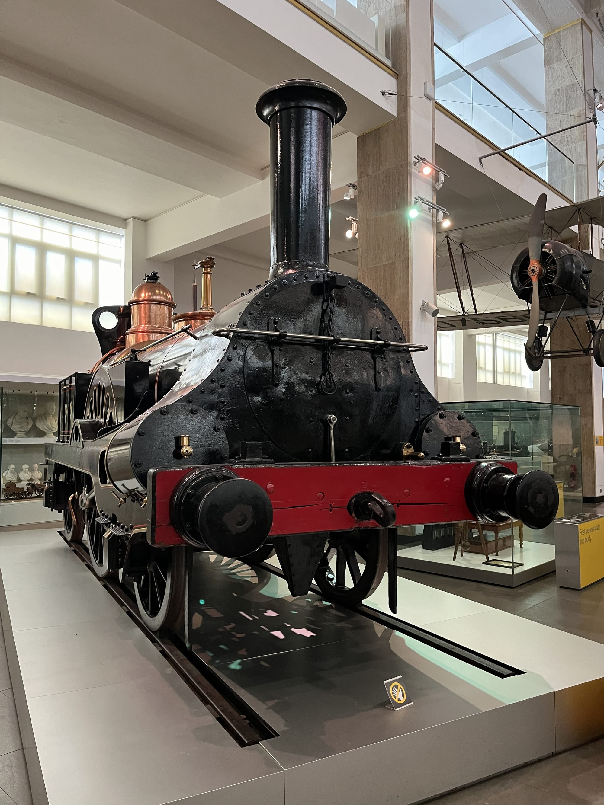 Historic steam locomotive displayed at the Science Museum in London, featuring a black boiler, tall chimney, copper fittings and red buffer beam, exhibited on a short section of railway track within the museum's transport gallery.