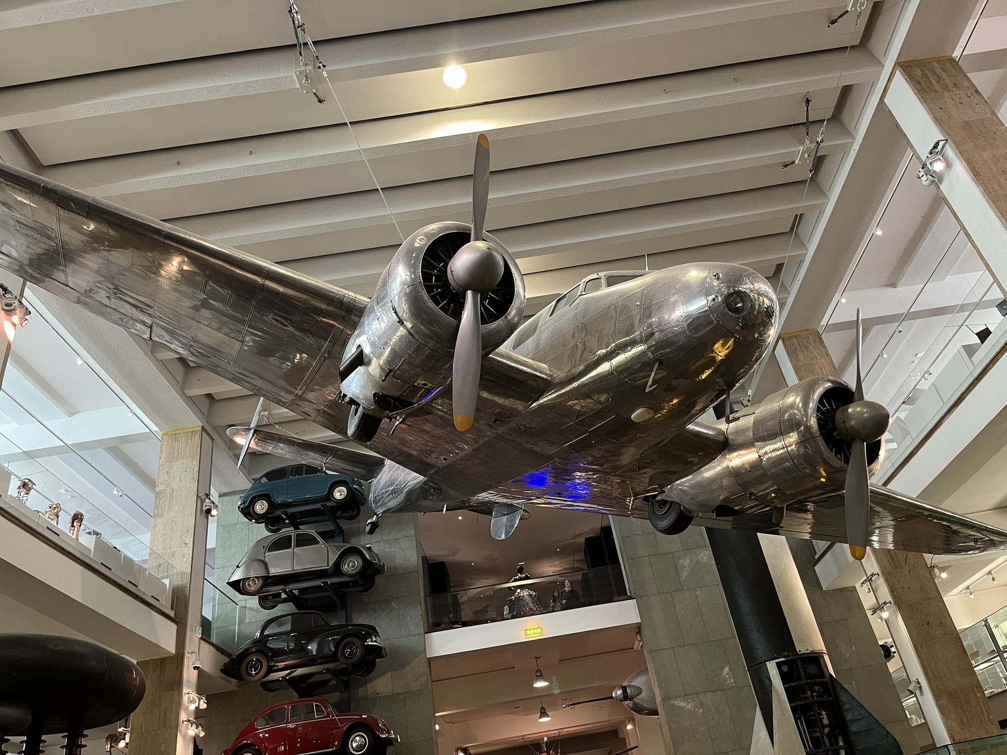 Historic twin‑engine propeller aircraft suspended from the ceiling in the Science Museum in London, viewed from below, with stacks of classic cars displayed nearby and transport exhibits filling the surrounding gallery.
