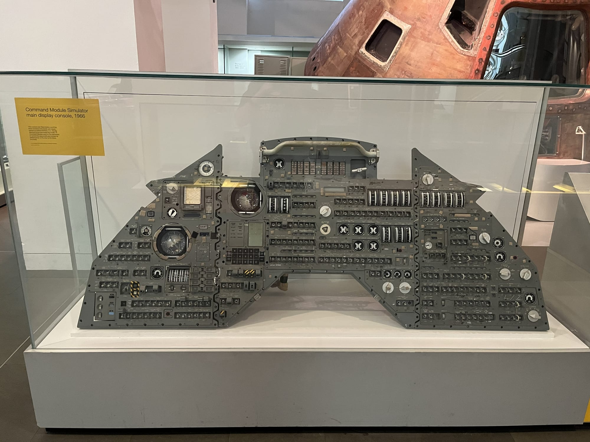 Command Module Simulator main display console from 1966 on display at the Science Museum in London, featuring a large panel of switches, dials, gauges and controls housed in a glass case, with an Apollo spacecraft capsule visible in the background.