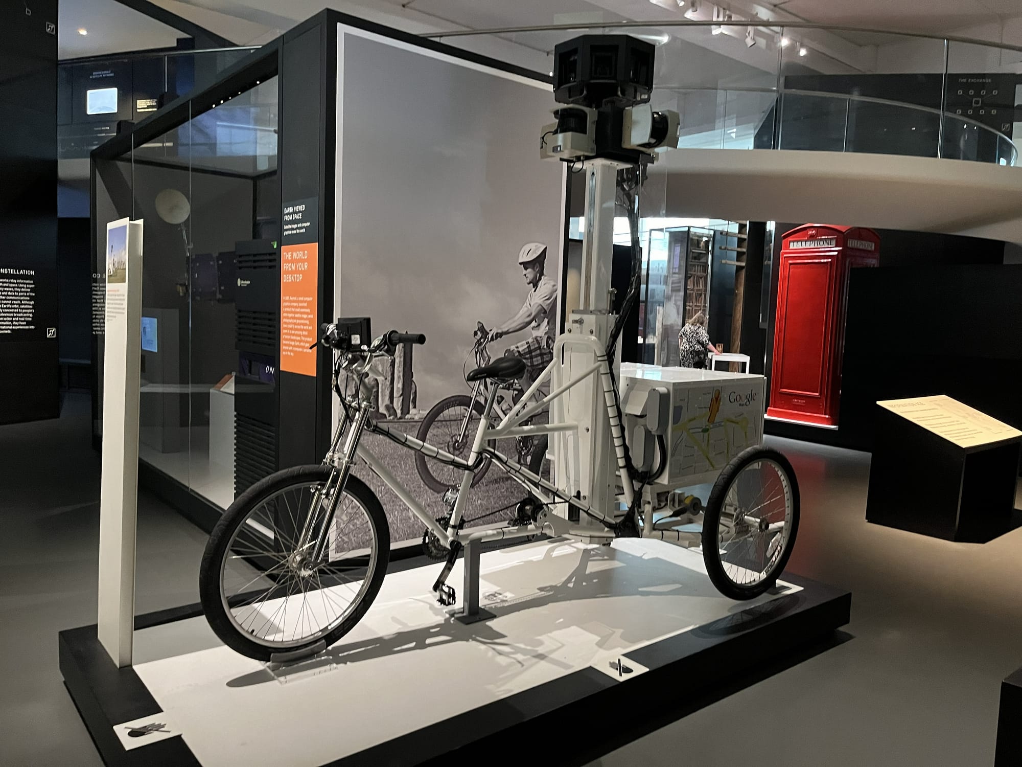 Google Street View tricycle on display at the Science Museum in London, featuring cameras mounted on a tall mast above a specially adapted three-wheeled bicycle used to capture panoramic imagery for Google Maps, with a red telephone box visible in the background.