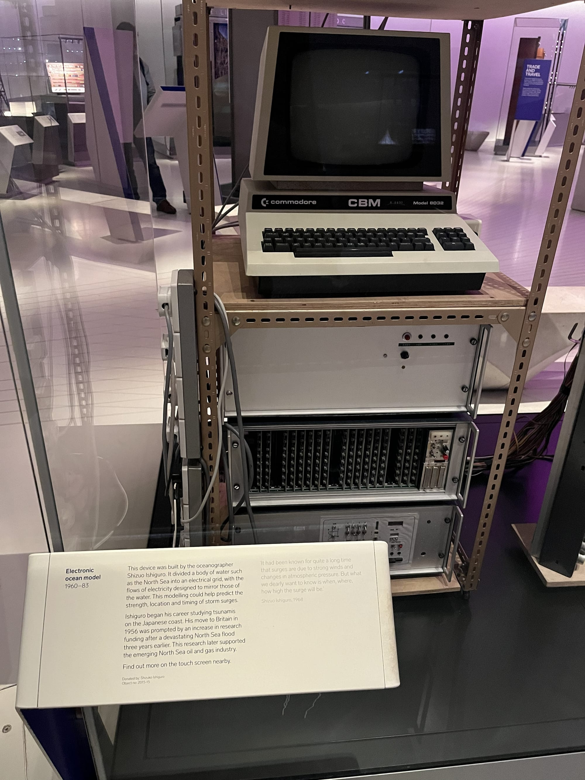 Electronic ocean model display at the Science Museum in London, featuring a Commodore CBM 8032 computer mounted above electronic equipment racks, displayed behind glass with an information panel describing its use in studying ocean currents and storm surges.
