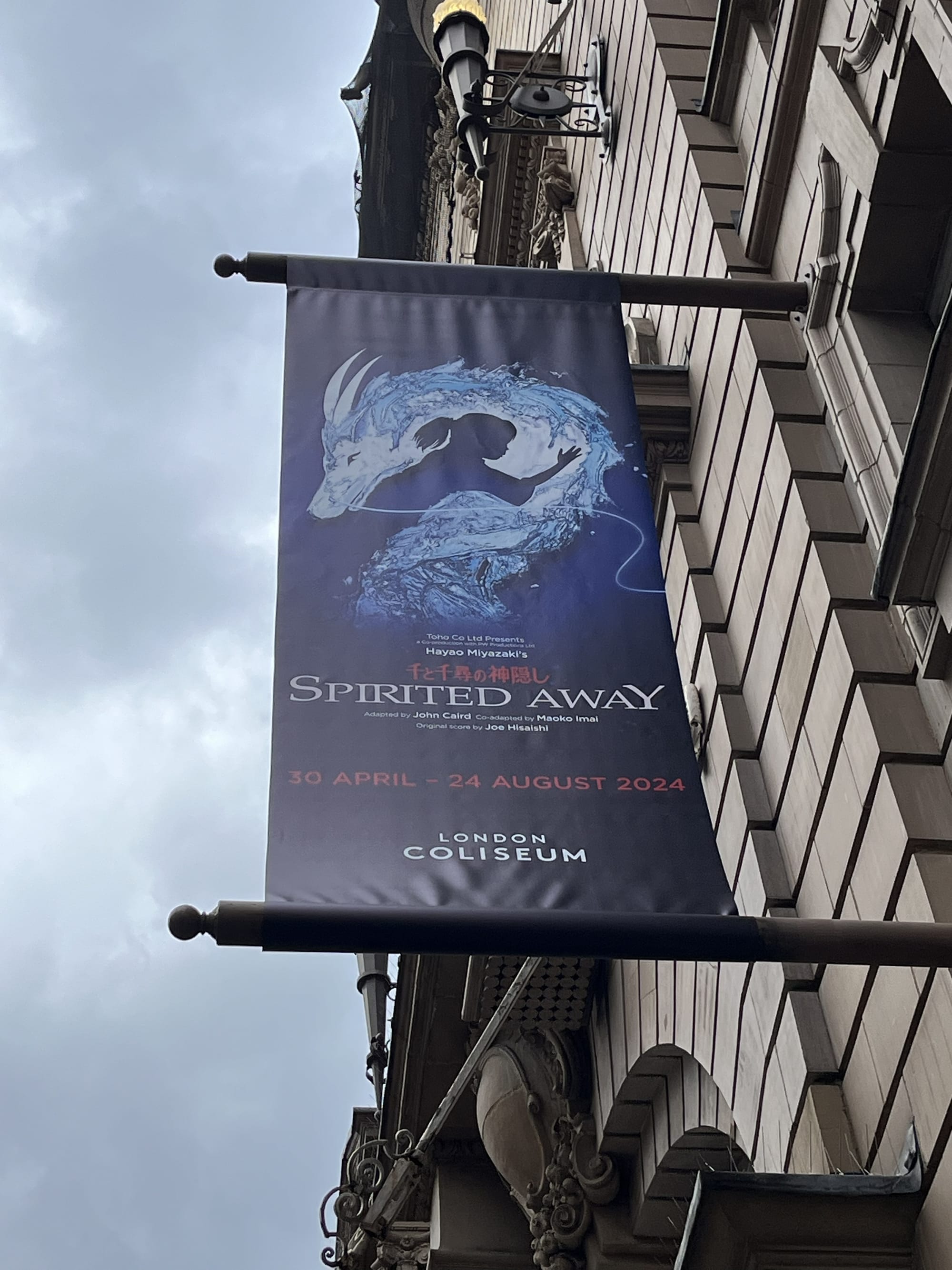 Banner advertising Spirited Away hanging outside the London Coliseum, featuring artwork inspired by Hayao Miyazaki's acclaimed story, with the theatre's ornate stone façade and a cloudy sky in the background.