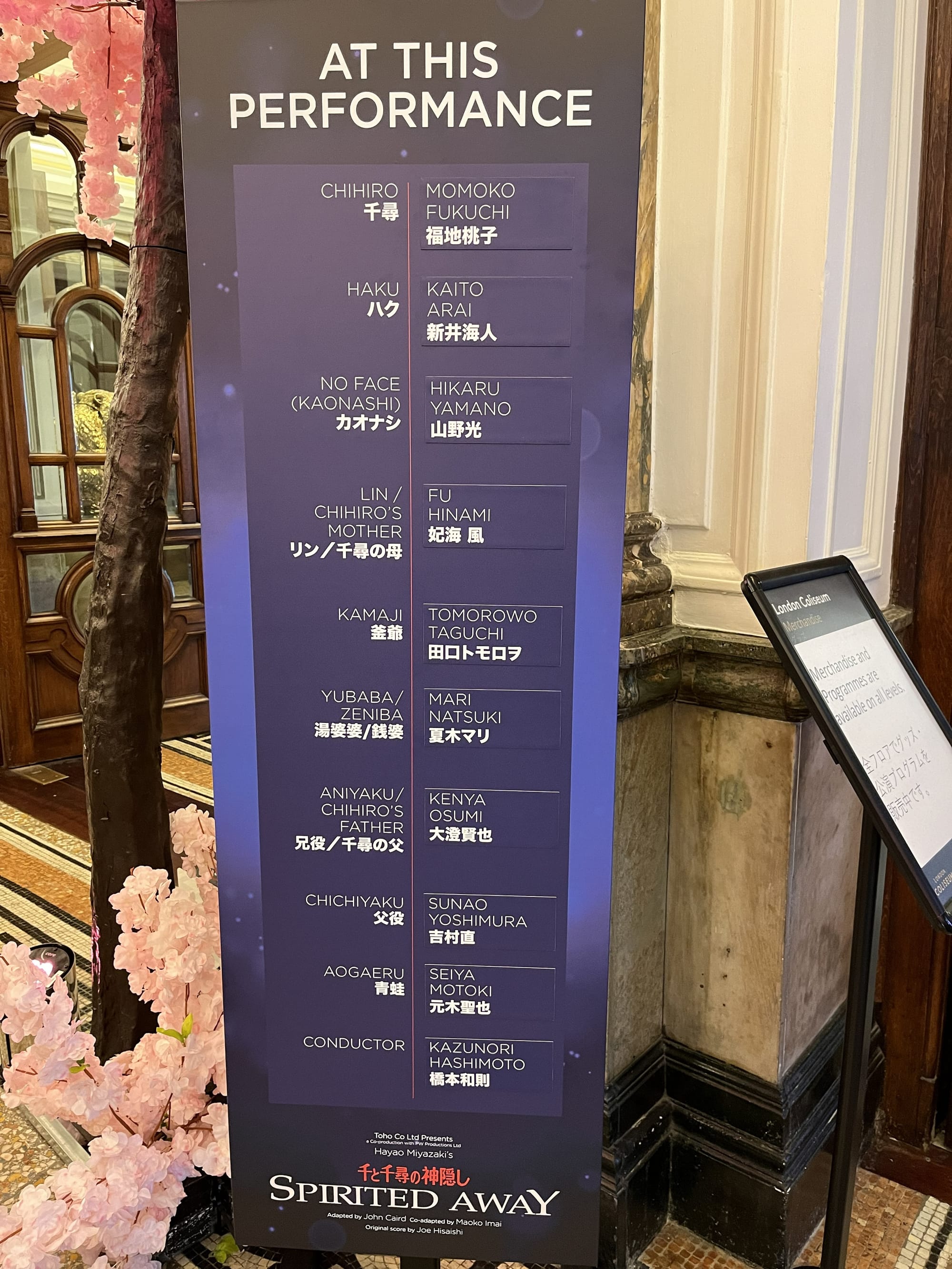Cast board for the stage production of Spirited Away at the London Coliseum, listing the performers appearing in that day's performance alongside character names in English and Japanese, displayed beside decorative cherry blossom theming in the theatre foyer.