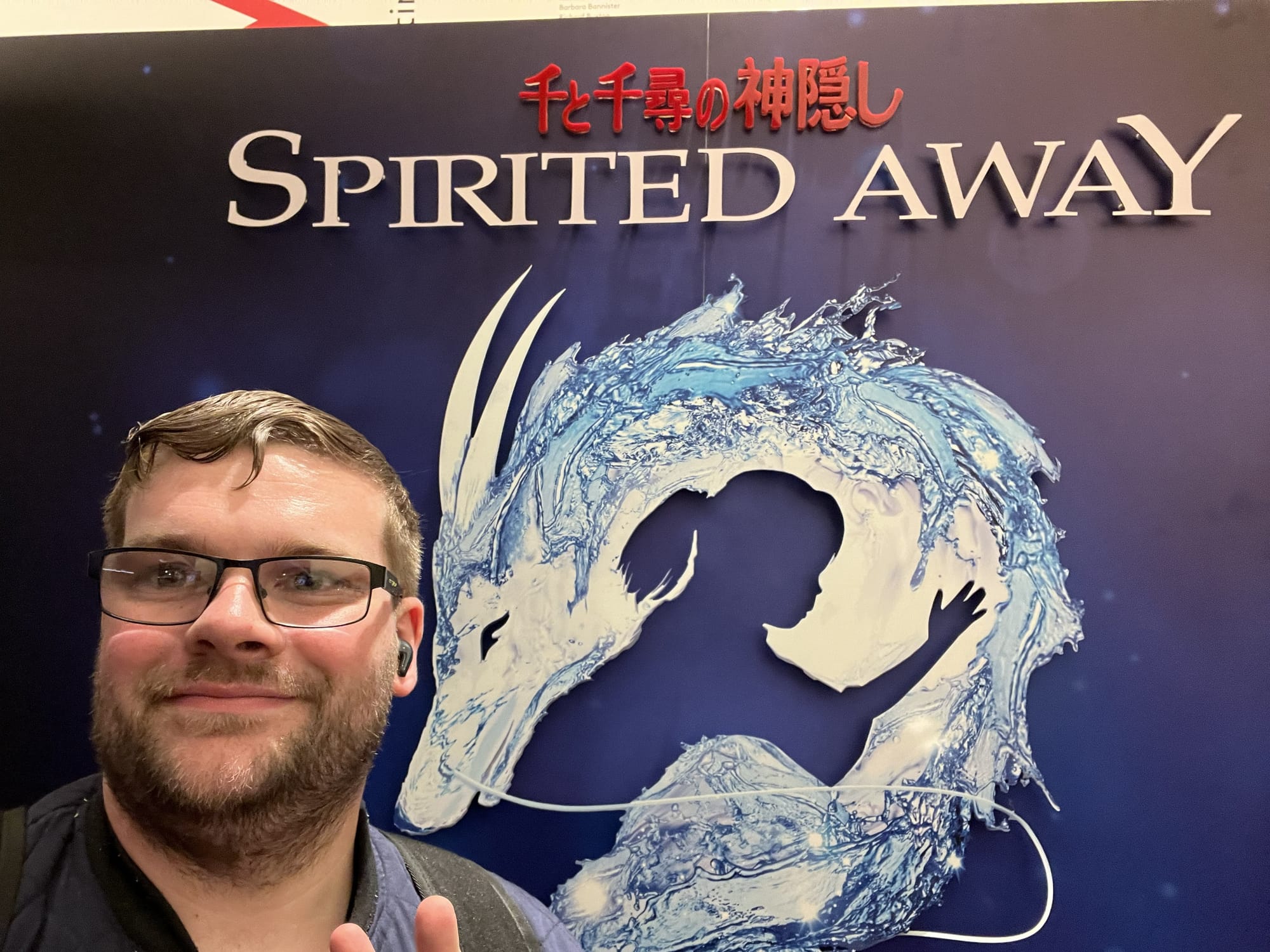 Visitor posing for a selfie in front of a Spirited Away promotional display, featuring the show's title and artwork inspired by the Studio Ghibli story, at the London Coliseum.