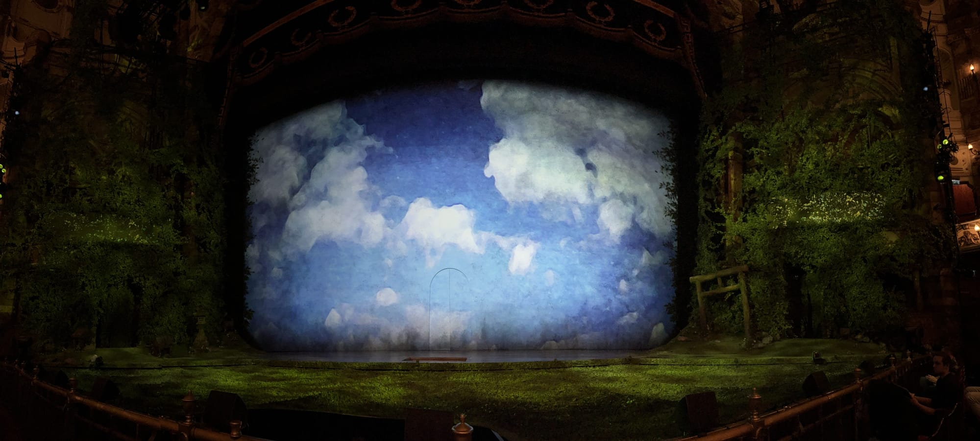 Stage set for Spirited Away at the London Coliseum, featuring a painted backdrop of blue sky and clouds framed by lush greenery, trees and a torii gate, creating a dreamlike landscape before the performance begins.