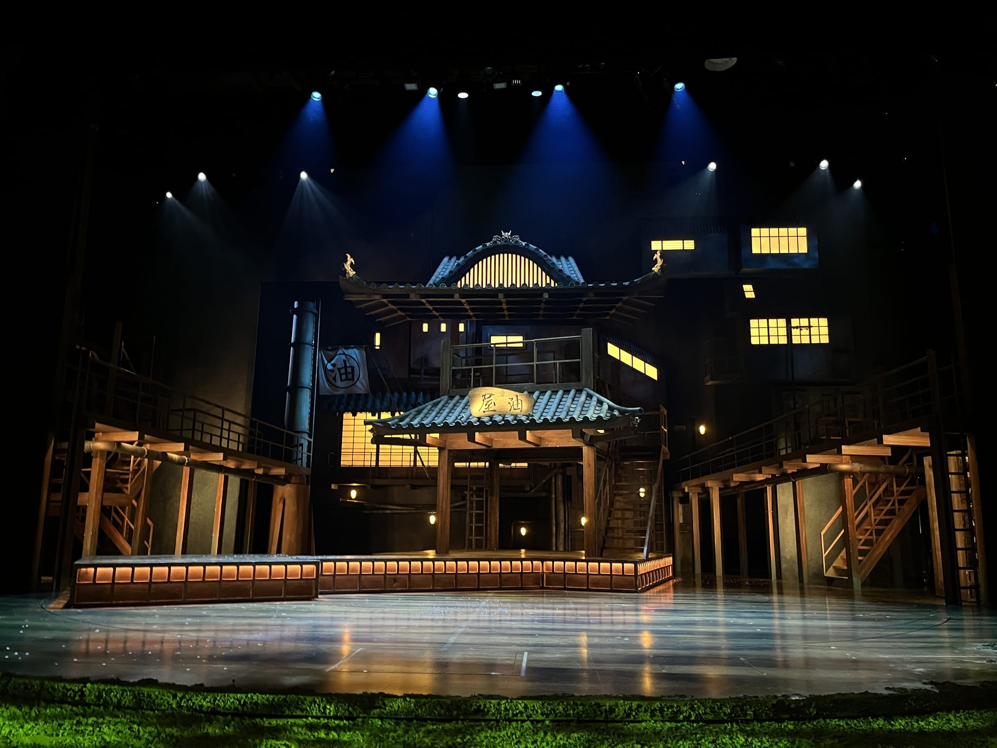 Stage set for Spirited Away at the London Coliseum, featuring a large traditional Japanese bathhouse with illuminated windows, wooden walkways and lantern-lit architecture beneath dramatic blue stage lighting.