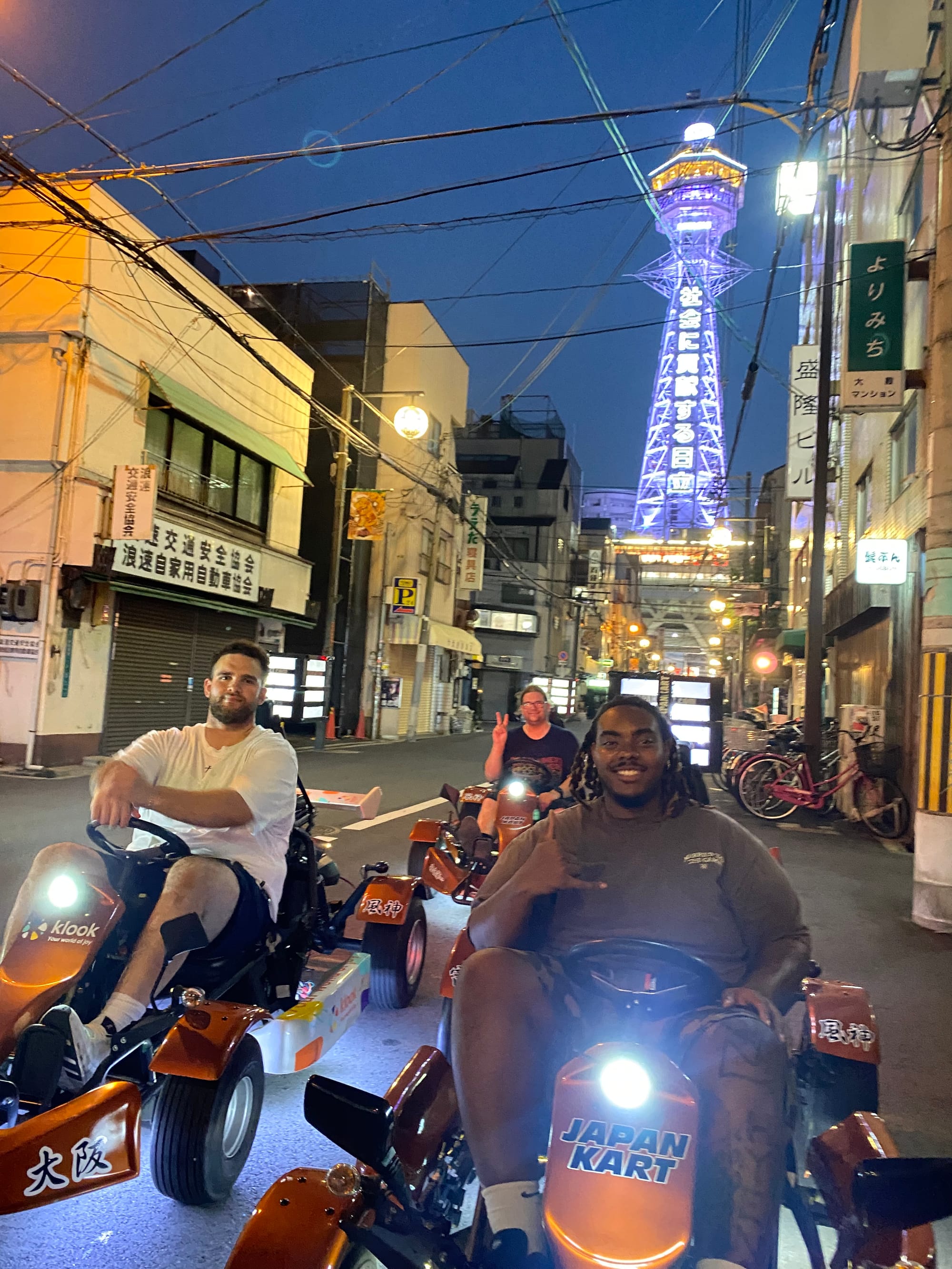 Participants drive go-karts through the streets of Osaka at night, with Tsutenkaku Tower illuminated in the background and city lights reflecting along the road.