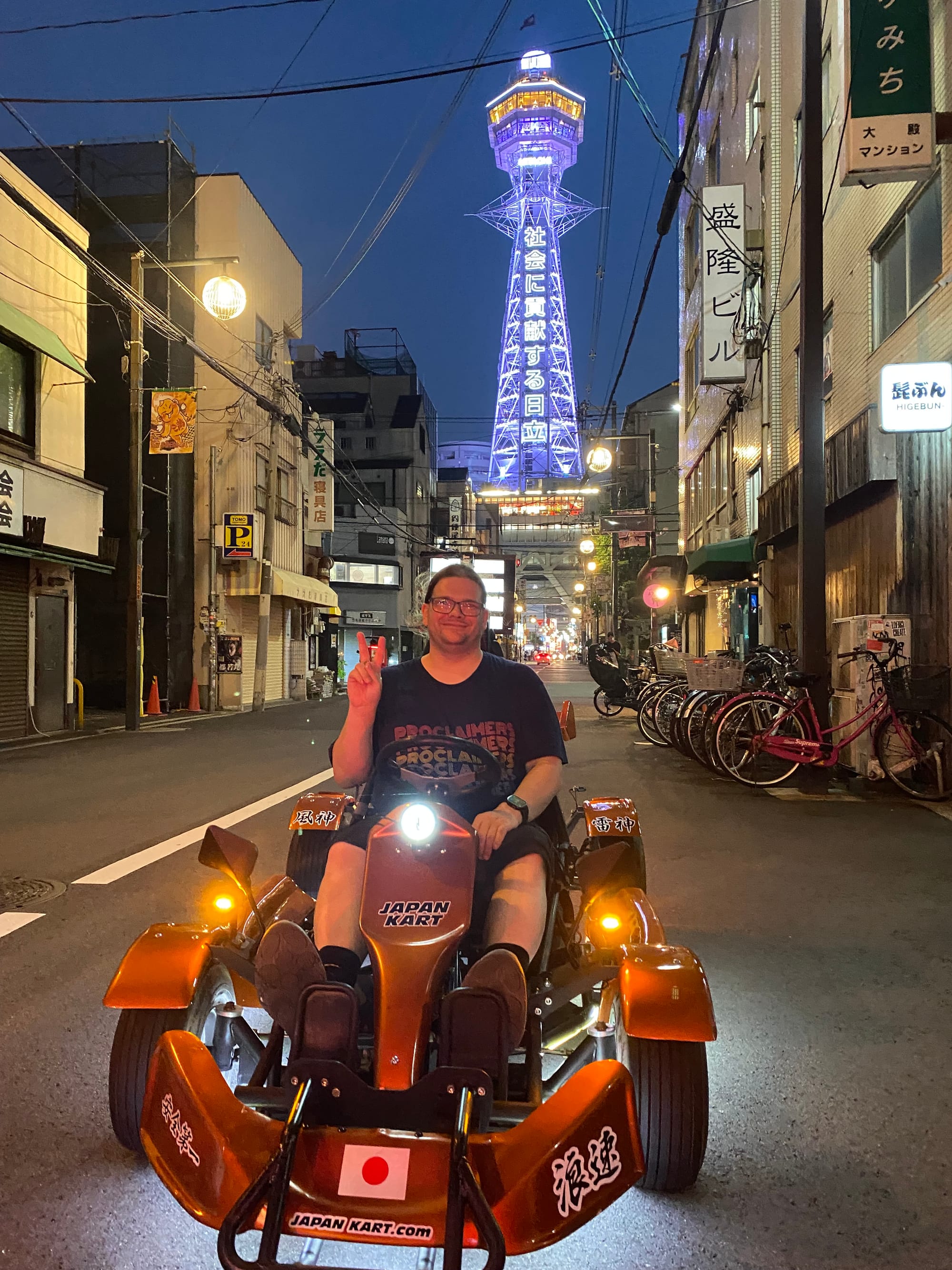 Visitor in a go-kart pauses on a street in Osaka's Shinsekai district at night, with the illuminated Tsutenkaku Tower rising above the surrounding buildings.
