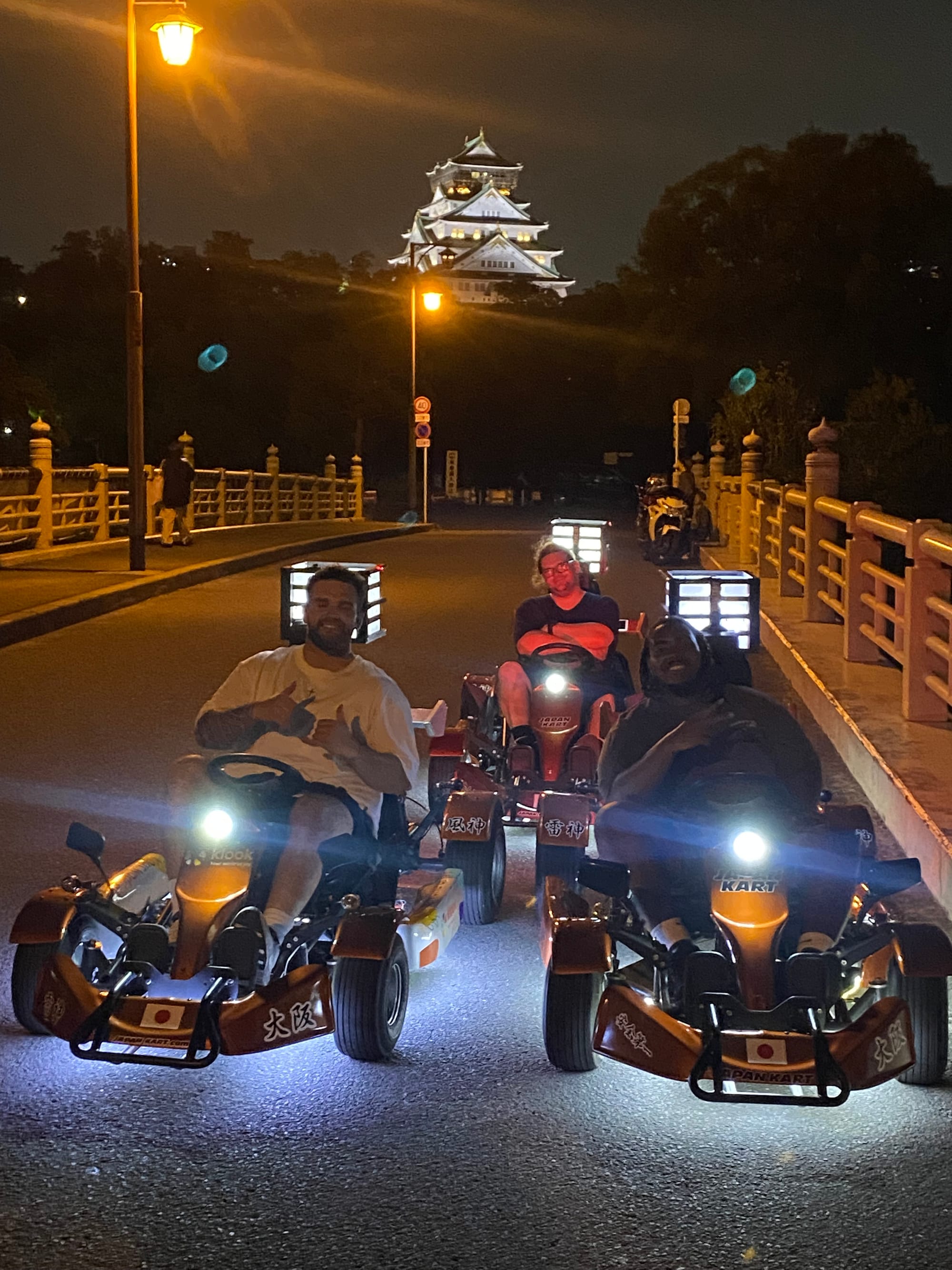 Go-karts parked on a bridge in Osaka at night, with Osaka Castle illuminated in the background and riders posing beneath the glow of street lamps.