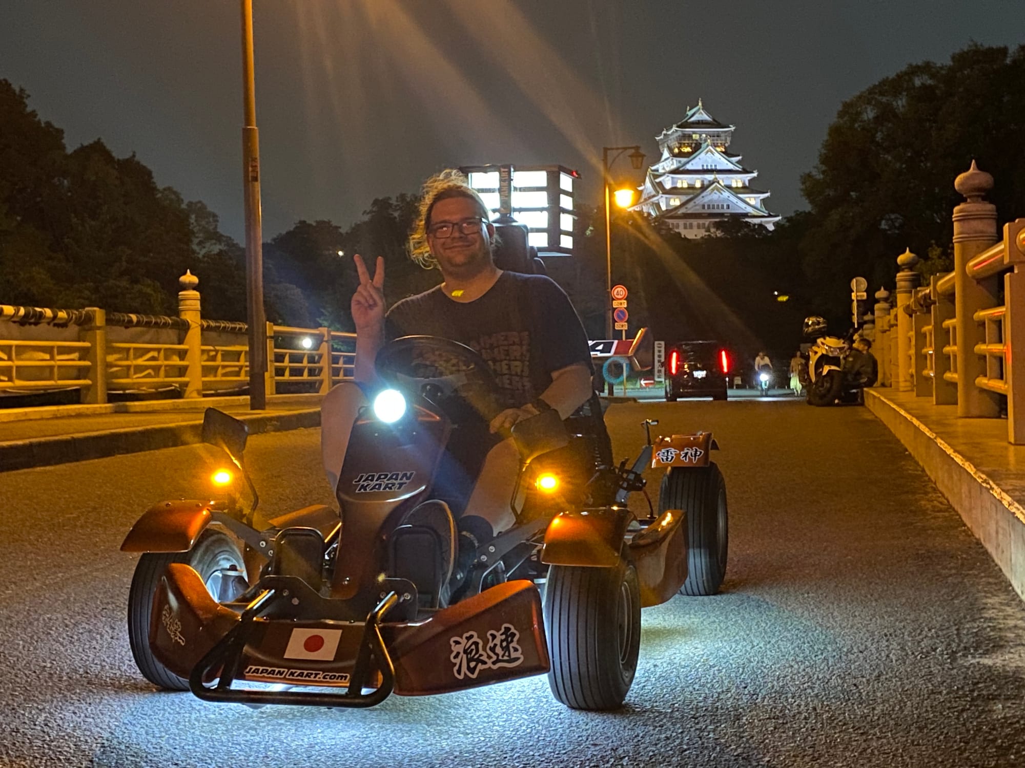 Visitor seated in a go-kart on a bridge in Osaka at night, making a peace sign, with the illuminated Osaka Castle rising above the trees in the background.