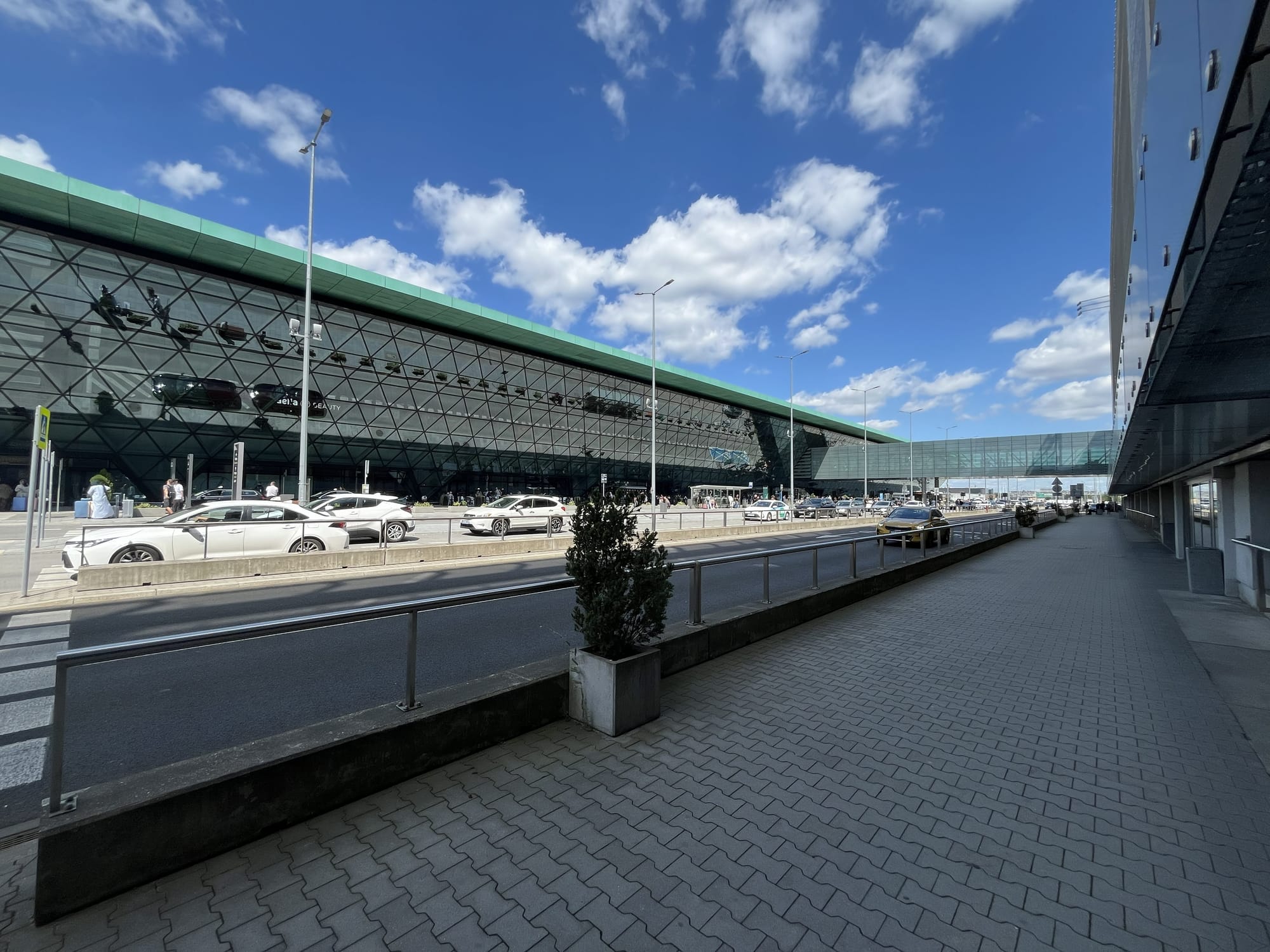 Exterior view of Kraków John Paul II International Airport, showing the modern glass-fronted terminal building, passenger drop-off area and vehicles under a bright blue sky with scattered clouds.