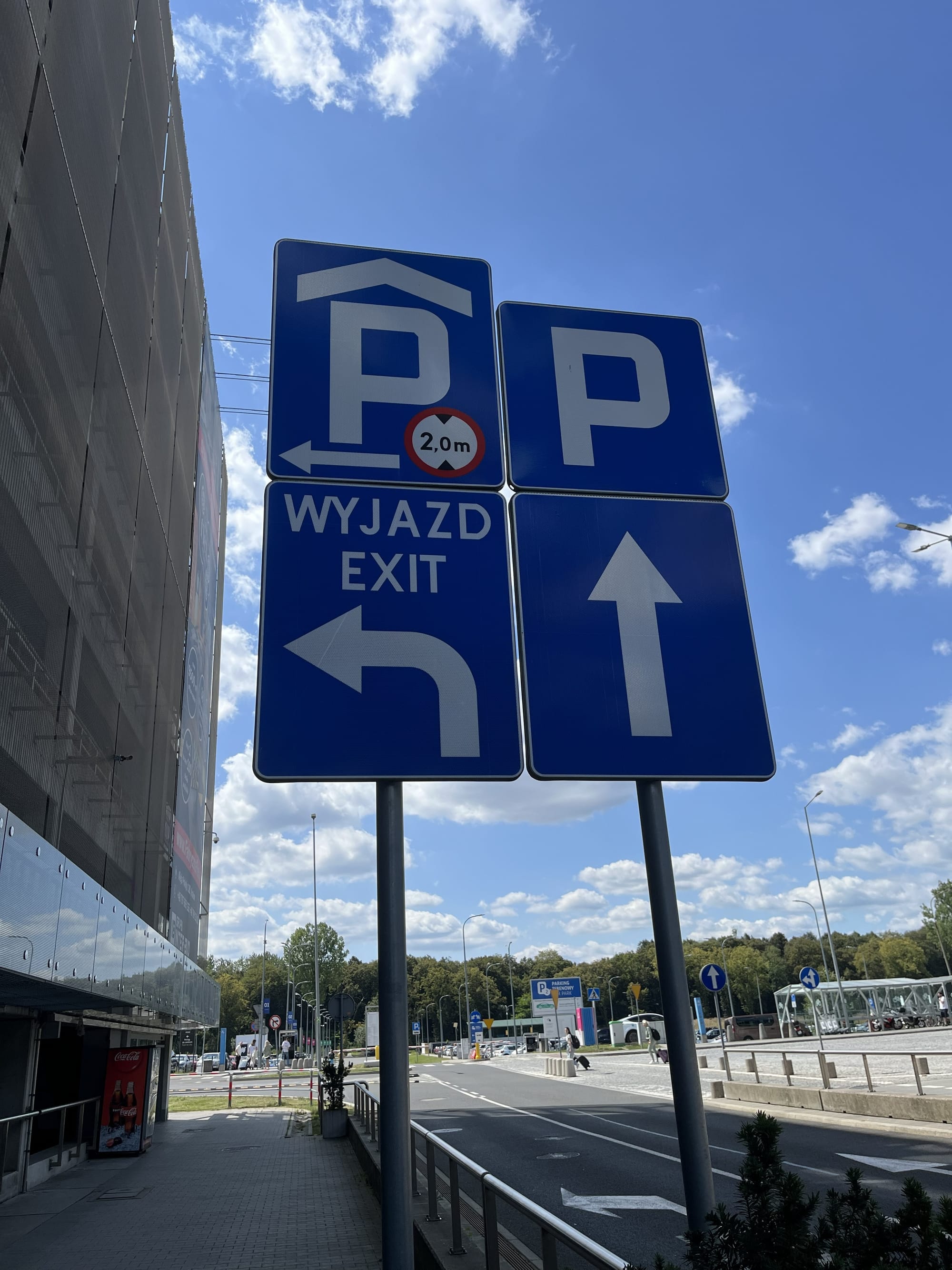 Blue parking and exit signs outside Kraków Airport, including bilingual Polish and English directions, with the terminal building and a bright blue sky in the background.
