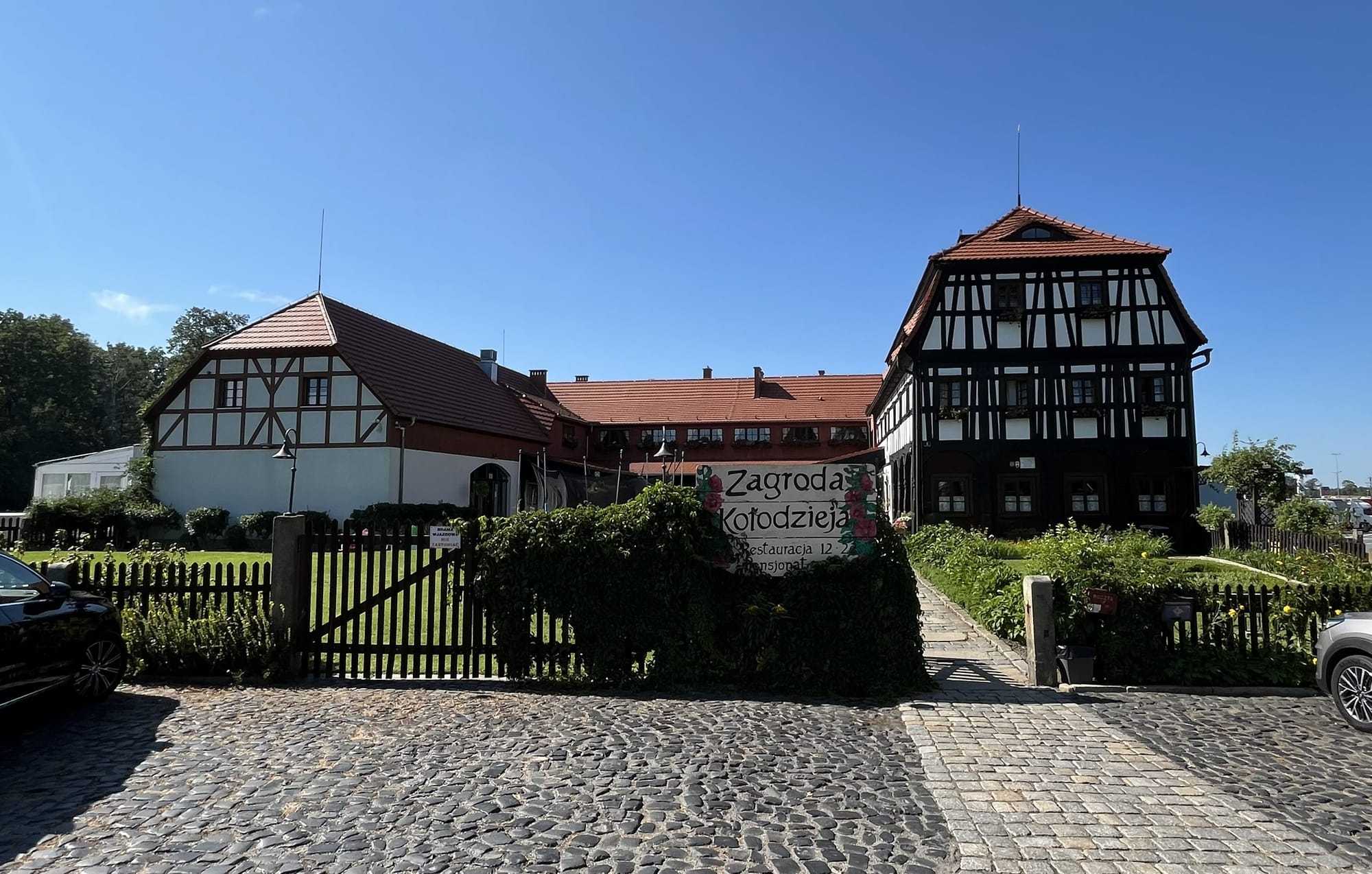 Traditional timber-framed buildings at Zagroda Kołodzieja in Zgorzelec, Poland, featuring historic Lusatian architecture, landscaped gardens and a sign for the hotel and restaurant beneath a clear blue sky.