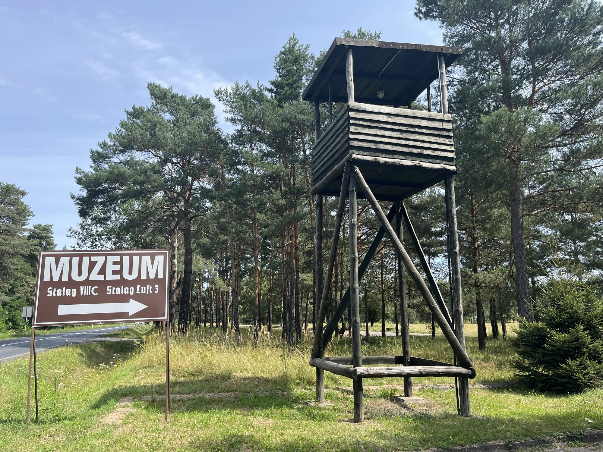 Replica wooden guard tower beside a museum sign for Stalag VIII C and Stalag Luft III, standing among pine trees near the site of the former prisoner-of-war camps.